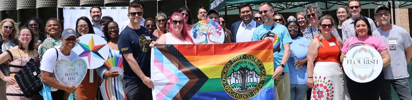 A group of Chicago Park District employees pose outdoors holding a pride flag and colorful signs.