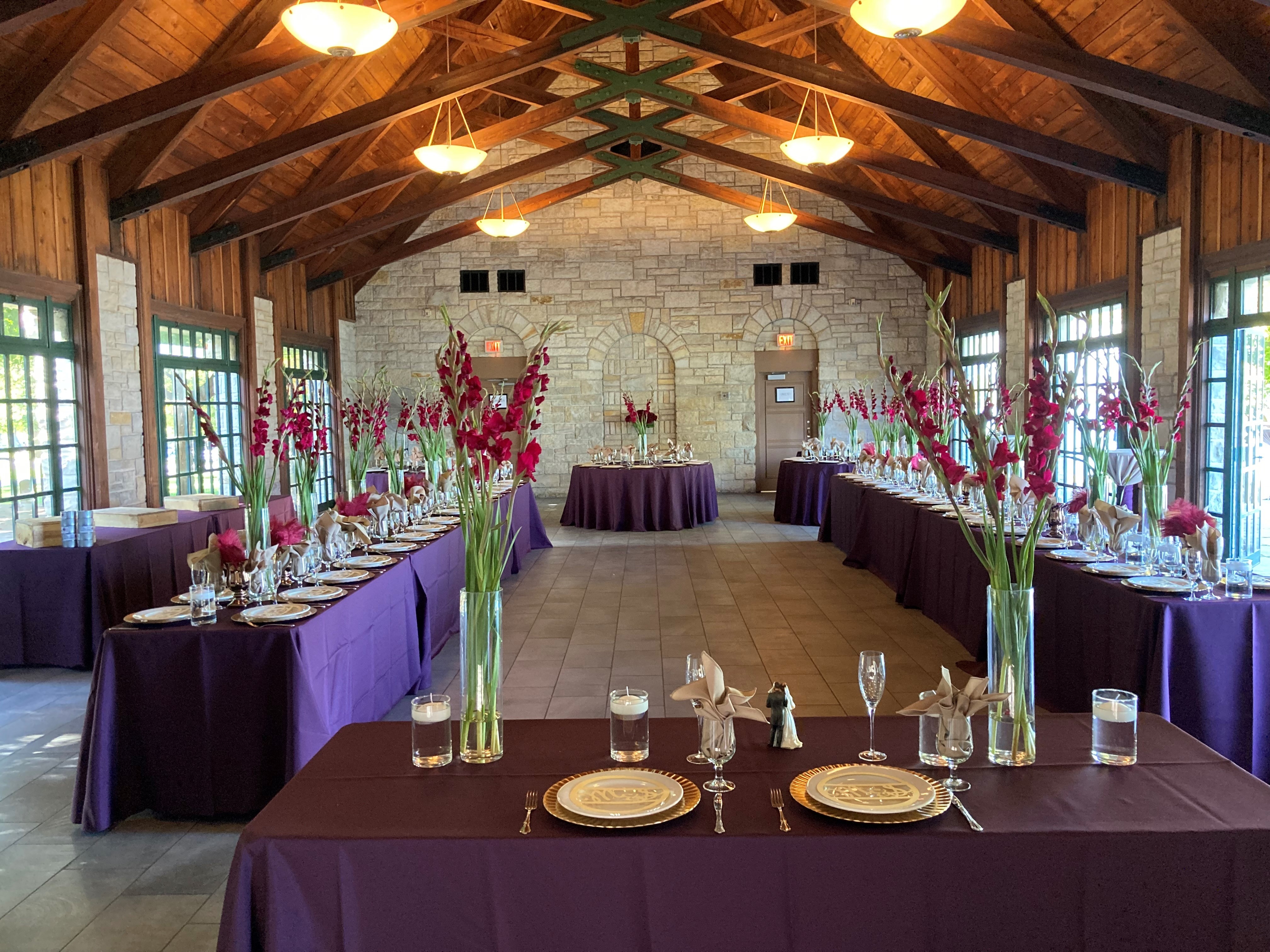 Wedding reception setup in a hall with purple tablecloths, floral centerpieces, and place settings.