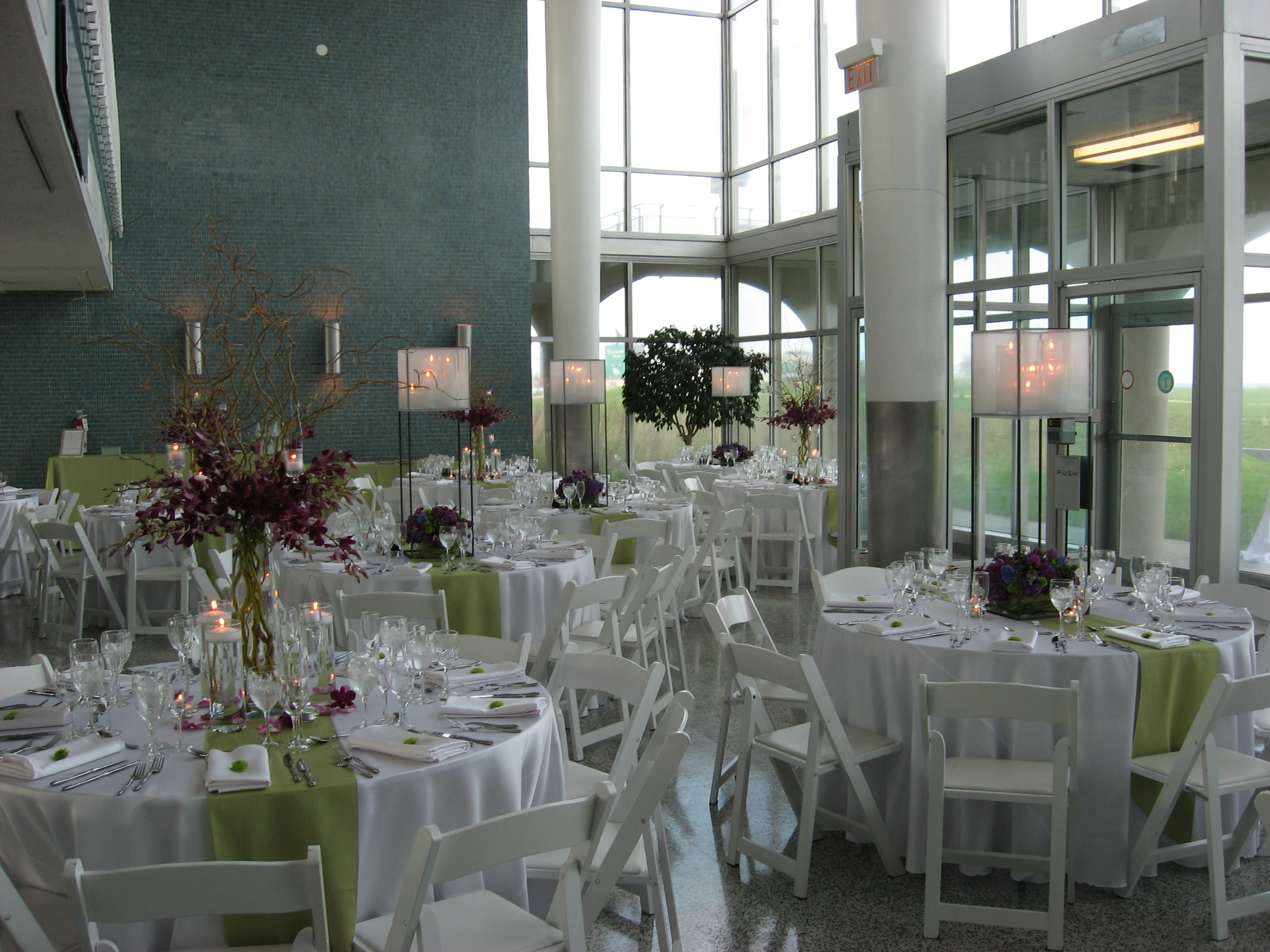 Elegant reception hall set for a wedding or banquet. Round tables with white and light green linens are surrounded by white folding chairs. Large floral centerpieces and tall candle holders decorate each table. Tall windows and teal tiled wall create a modern ambiance.