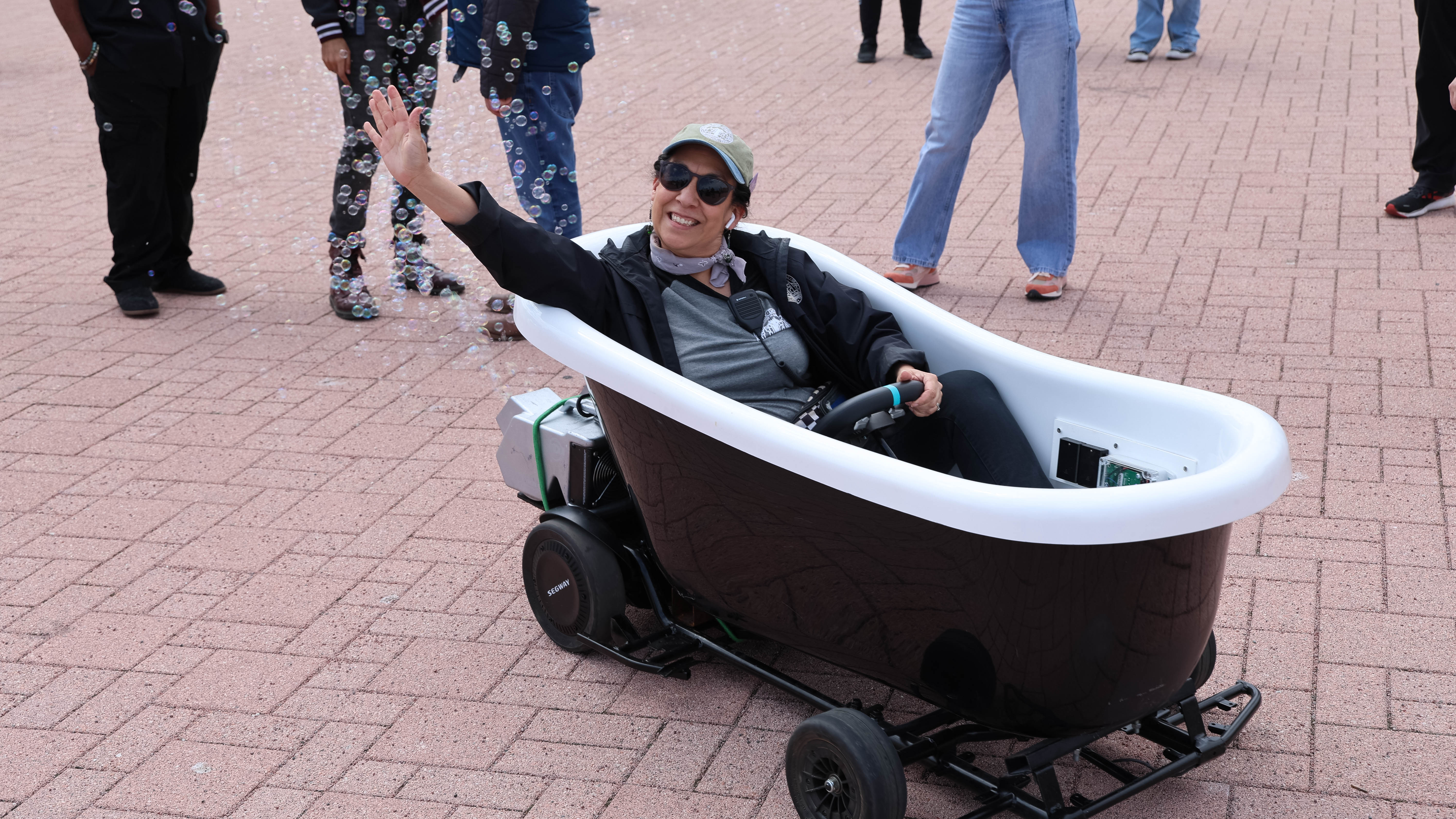 Woman waves while driving a motorized bathtub outdoors surrounded by bubbles.