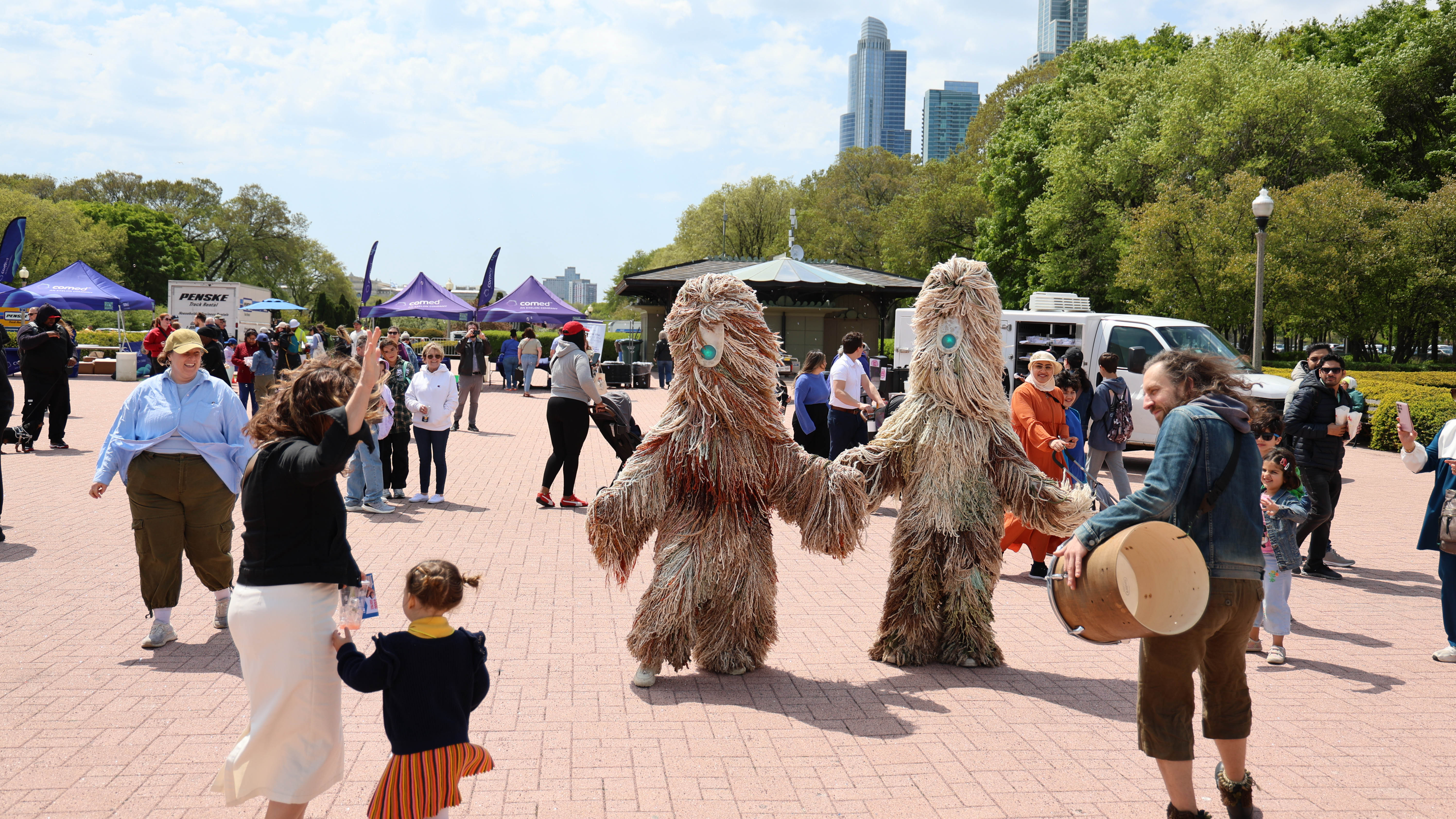 Two people in shaggy costumes hold hands as a drummer plays nearby at an outdoor festival.