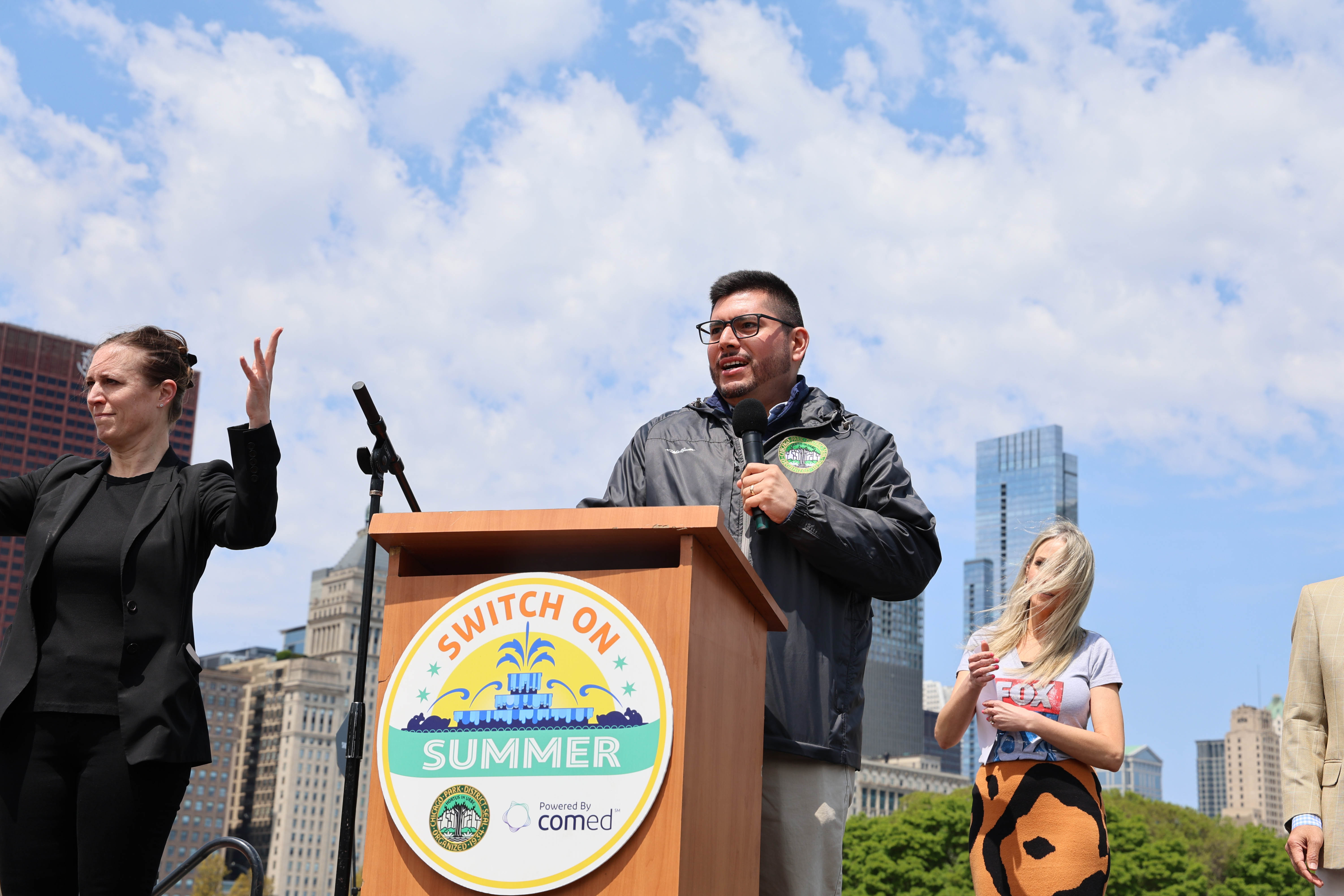 Man speaks at podium, flanked by ASL interpreters, with Chicago skyline in background.