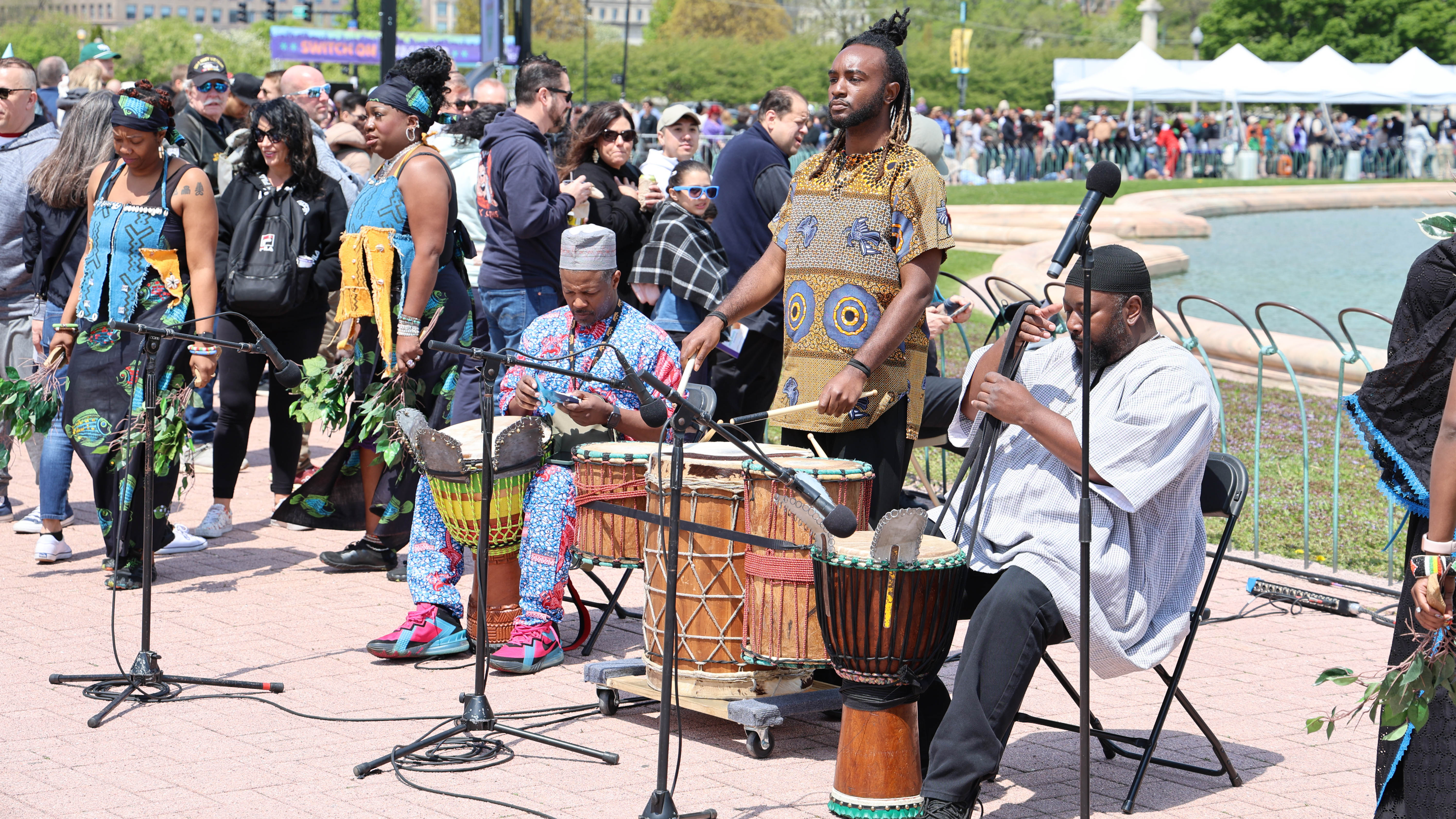Three drummers play traditional African drums at an outdoor festival. A crowd watches in the background.