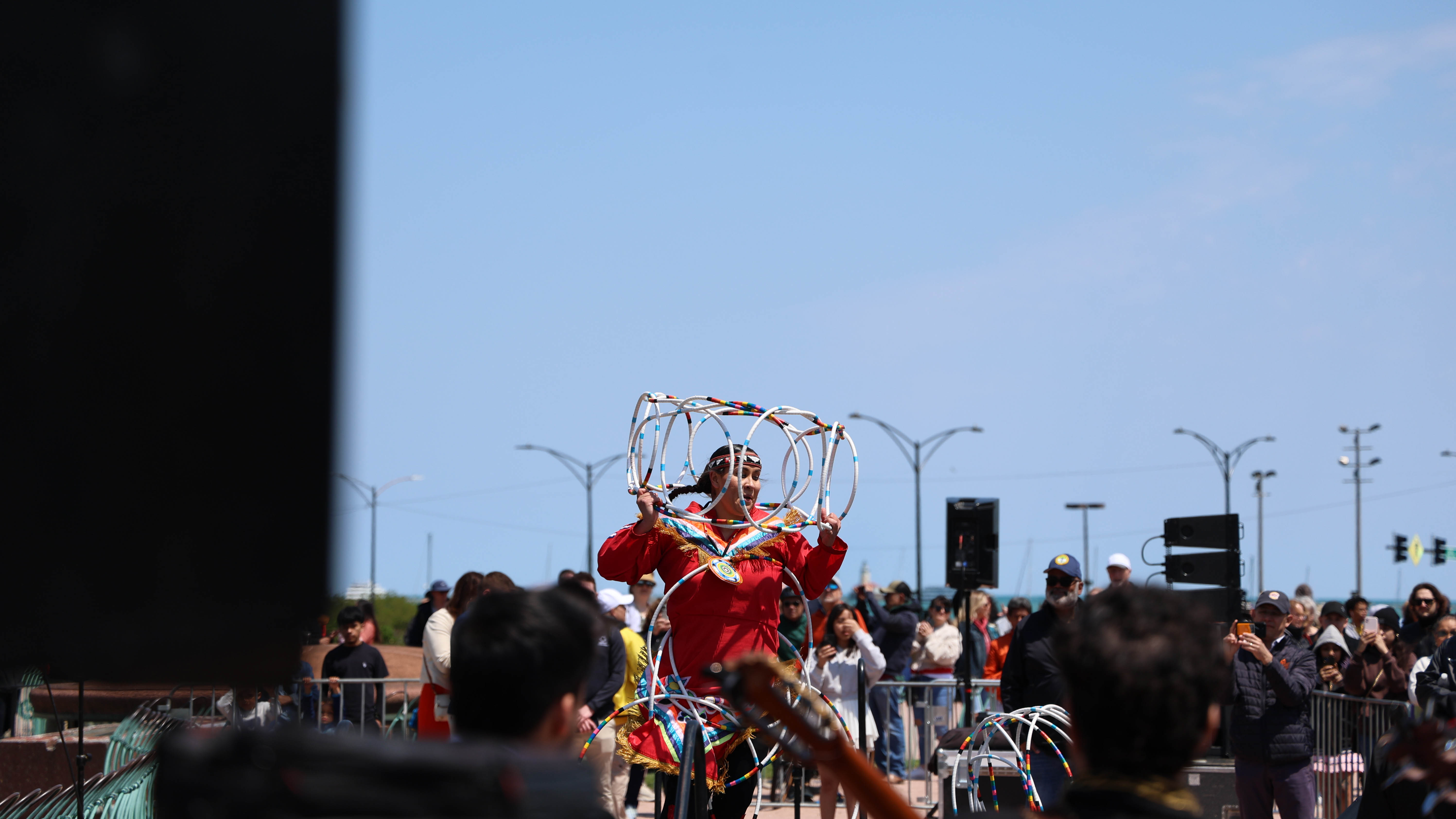 Indigenous dancer in red dress manipulates large hoops on a sunny outdoor stage. Crowd watches and takes photos.