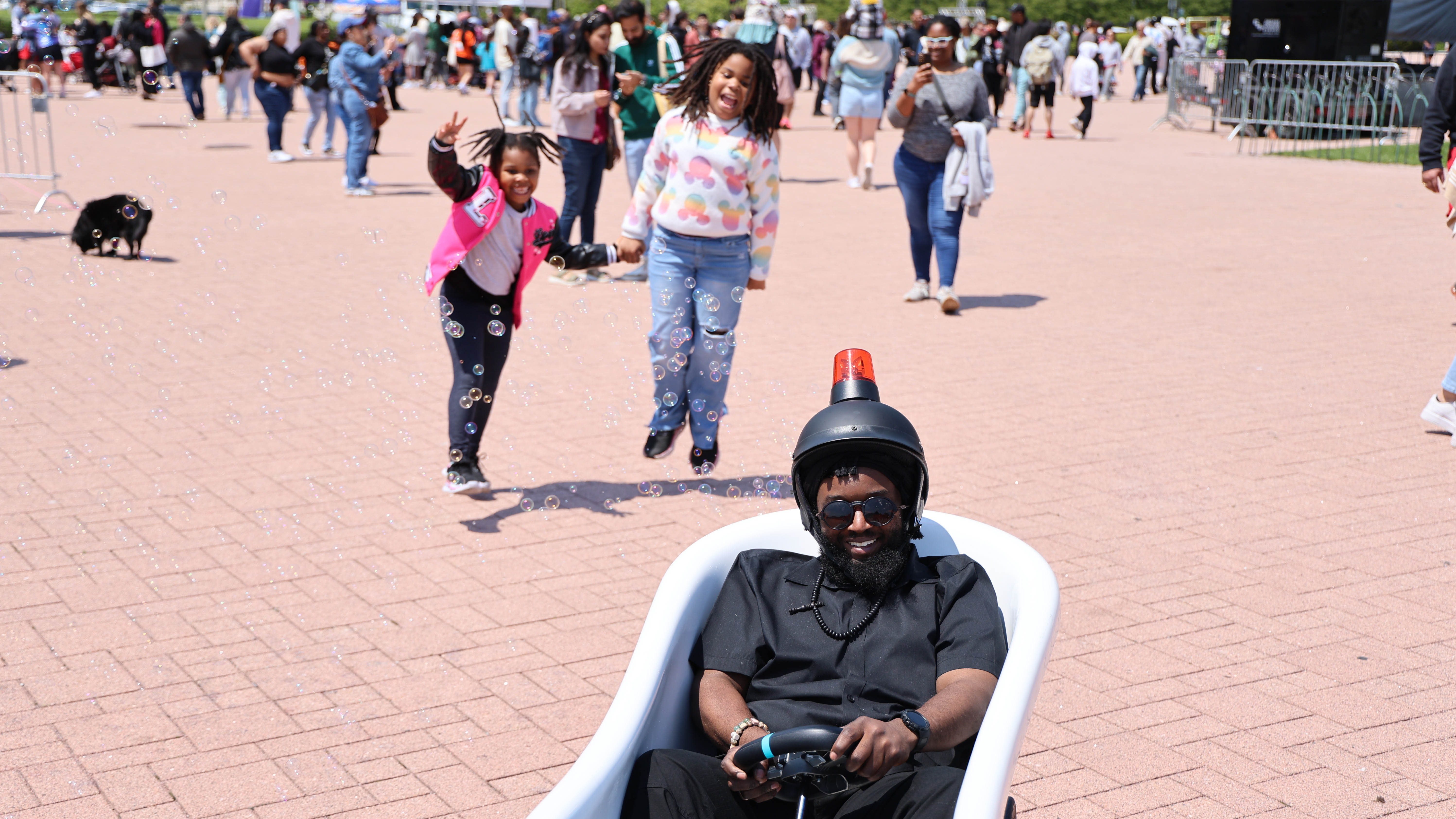 Man in a rolling bathtub smiles as two children chase bubbles.