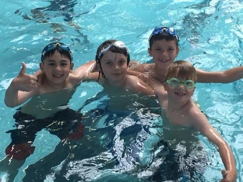 Four children wearing goggles smile and play together in a swimming pool. One gives a thumbs up.