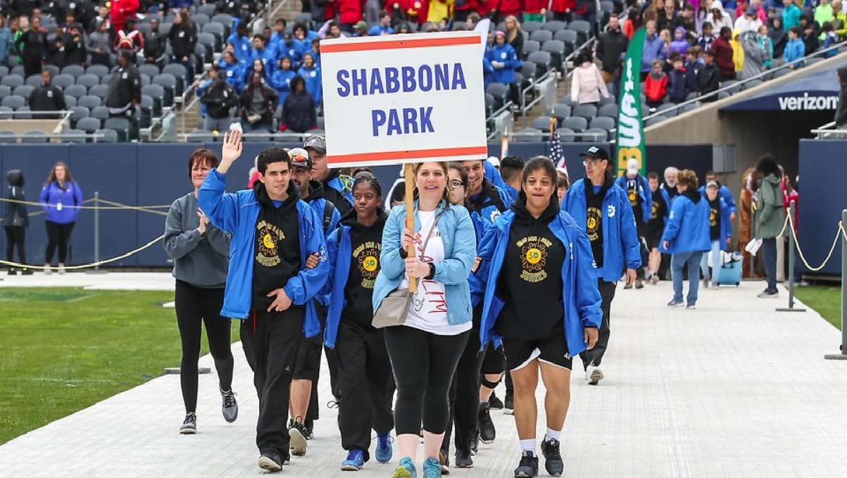 Group walks across stadium track carrying "Shabbona Park" sign.