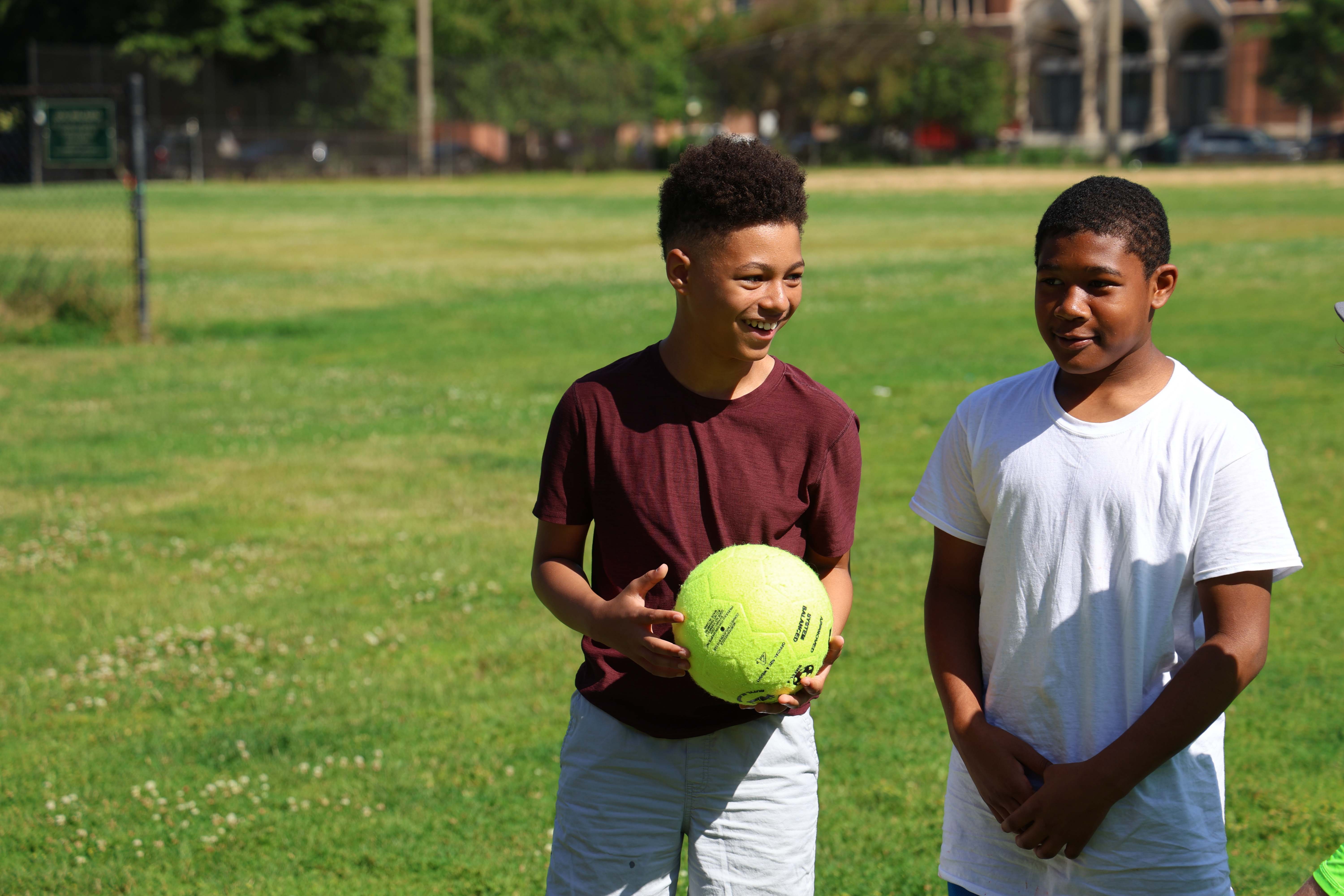 Two boys on a grassy field, one holding a neon yellow soccer ball.