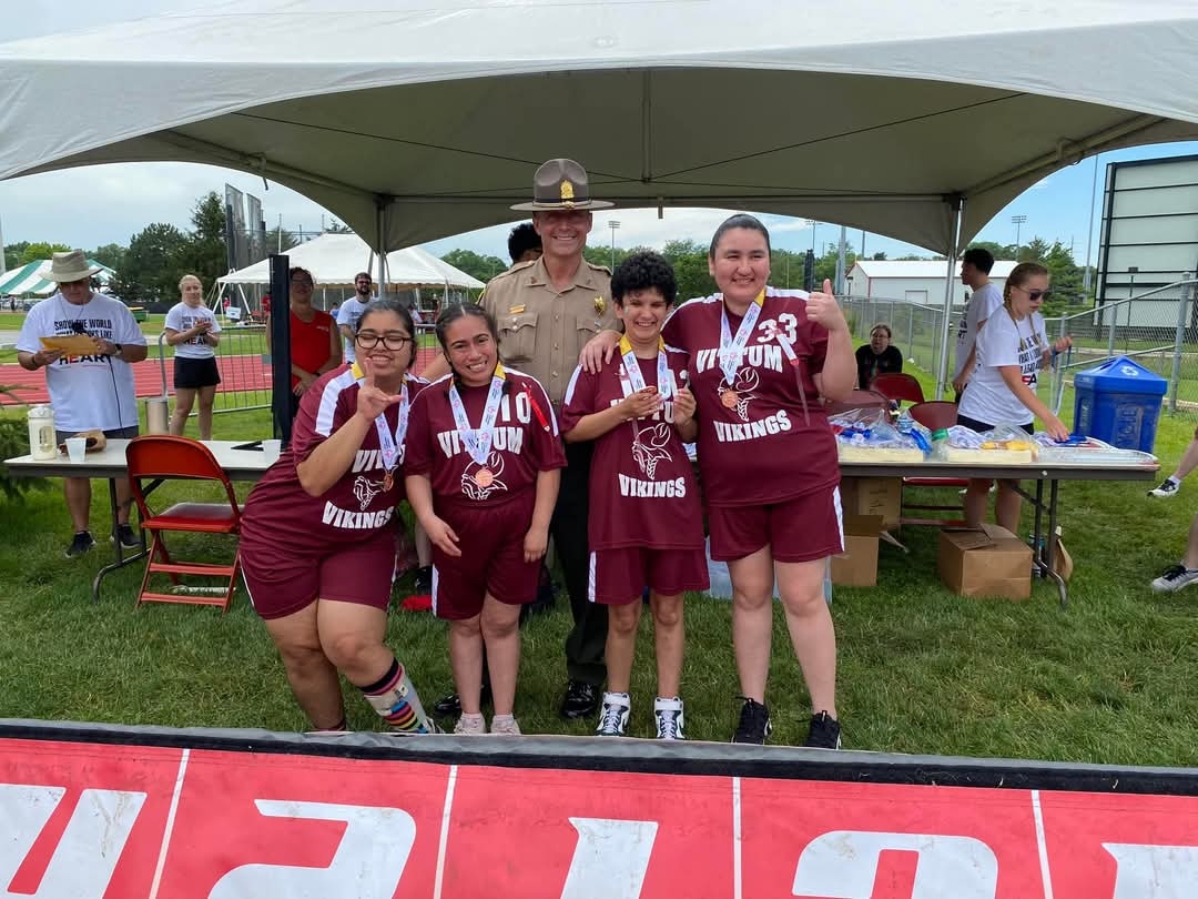 Three athletes with medals pose with a state trooper under a tent at a track and field event.