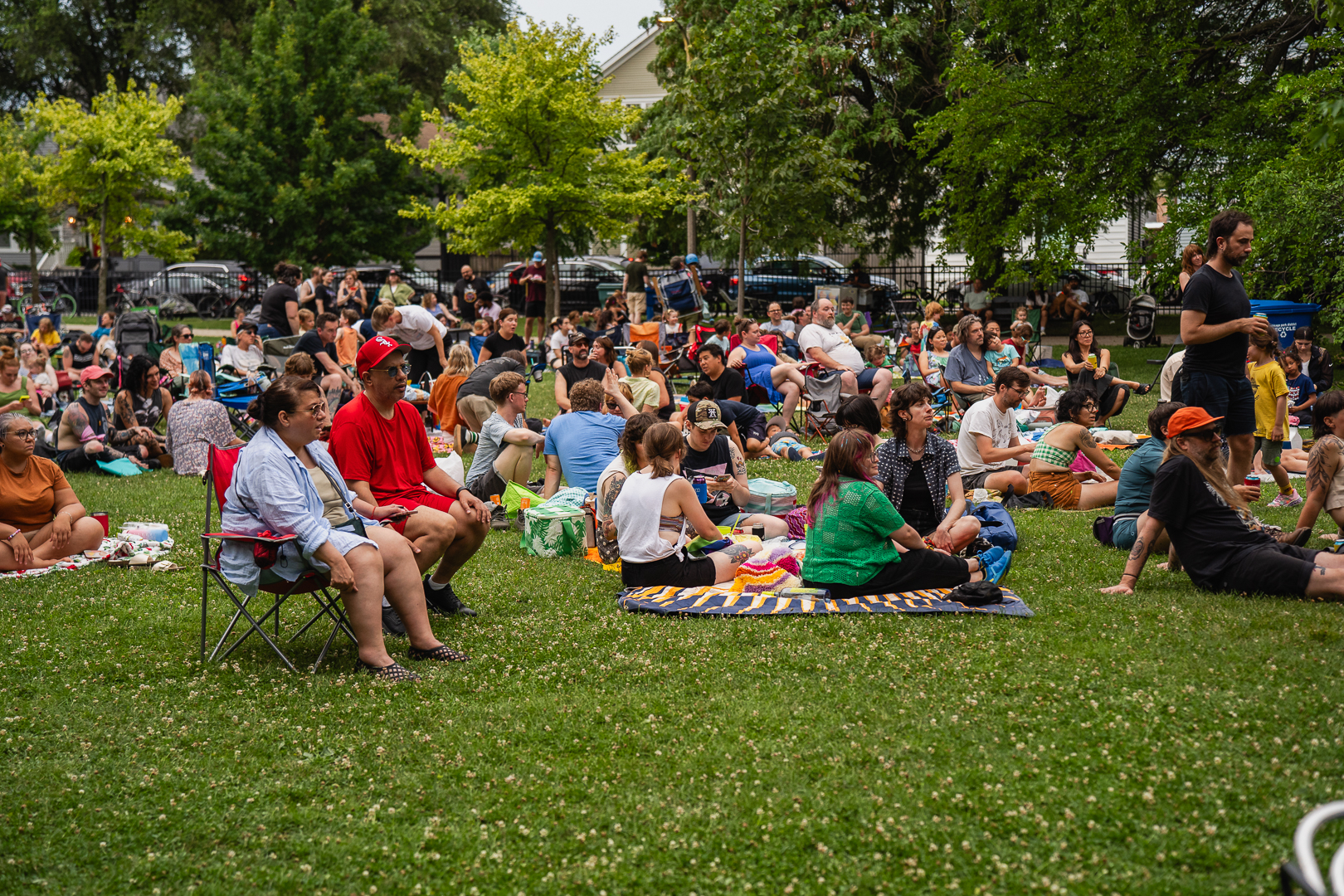 A crowd of people sit on blankets and lawn chairs in a grassy park.