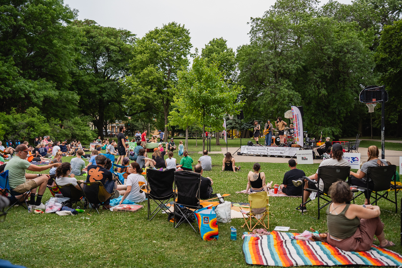 Crowd sits on lawn chairs and blankets in a park, watching a band play on a stage.