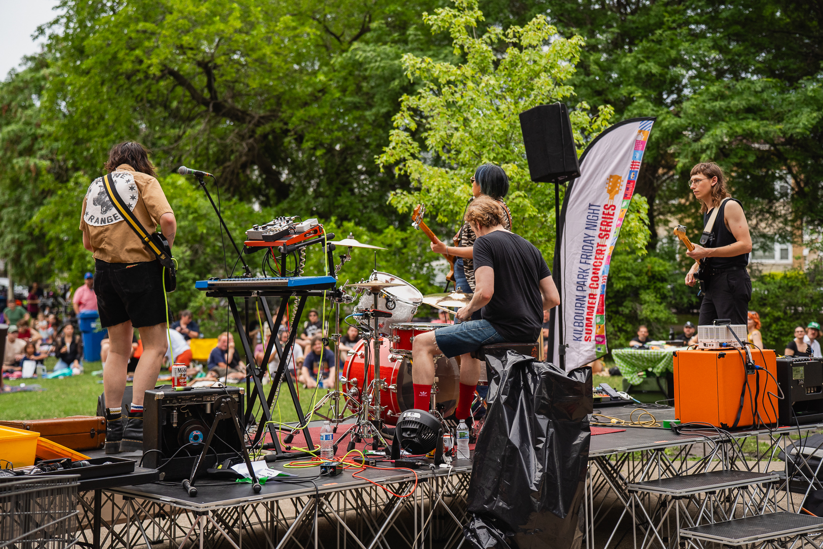 A band plays on an outdoor stage for an audience sitting on the grass.
