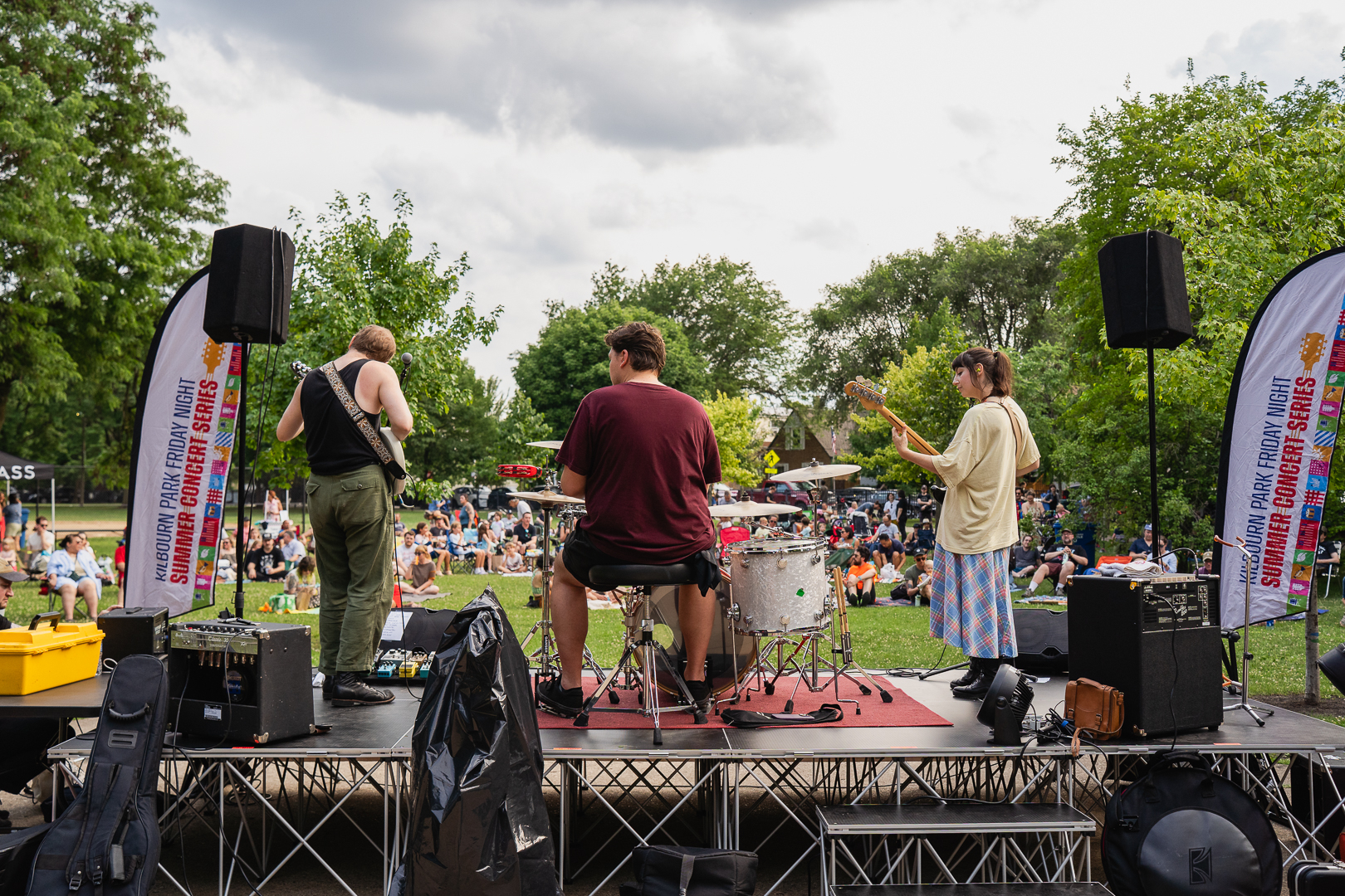A three-person band plays on an outdoor stage, their backs to the camera, facing a crowd seated on the grass.