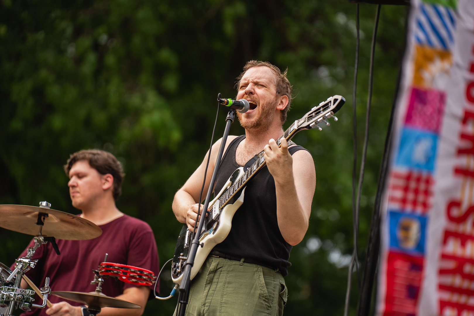 Musician sings into a microphone and plays a white electric guitar. A drummer is visible in the background.