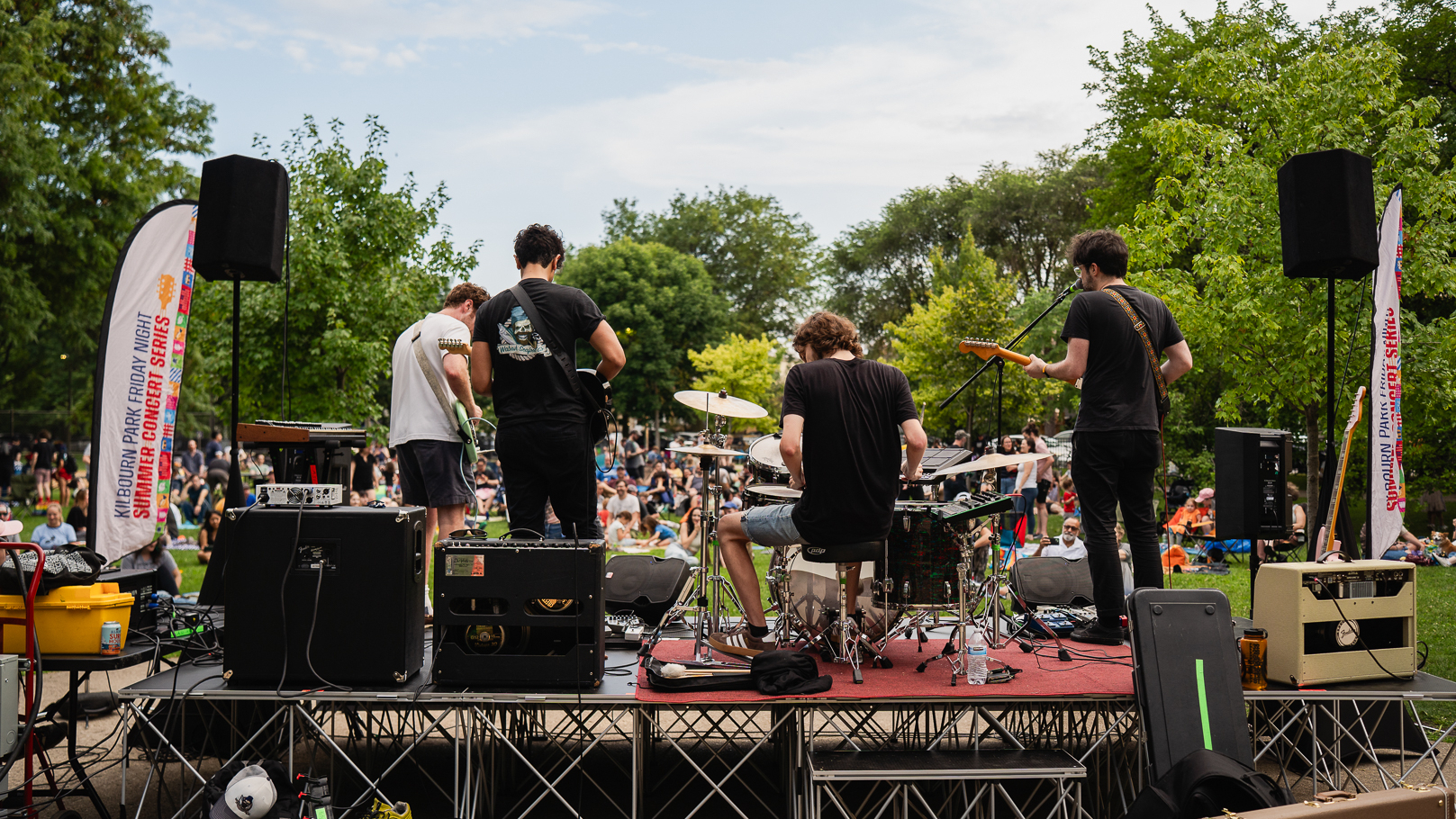 A band plays on an outdoor stage, seen from backstage, with the audience seated on the grass.