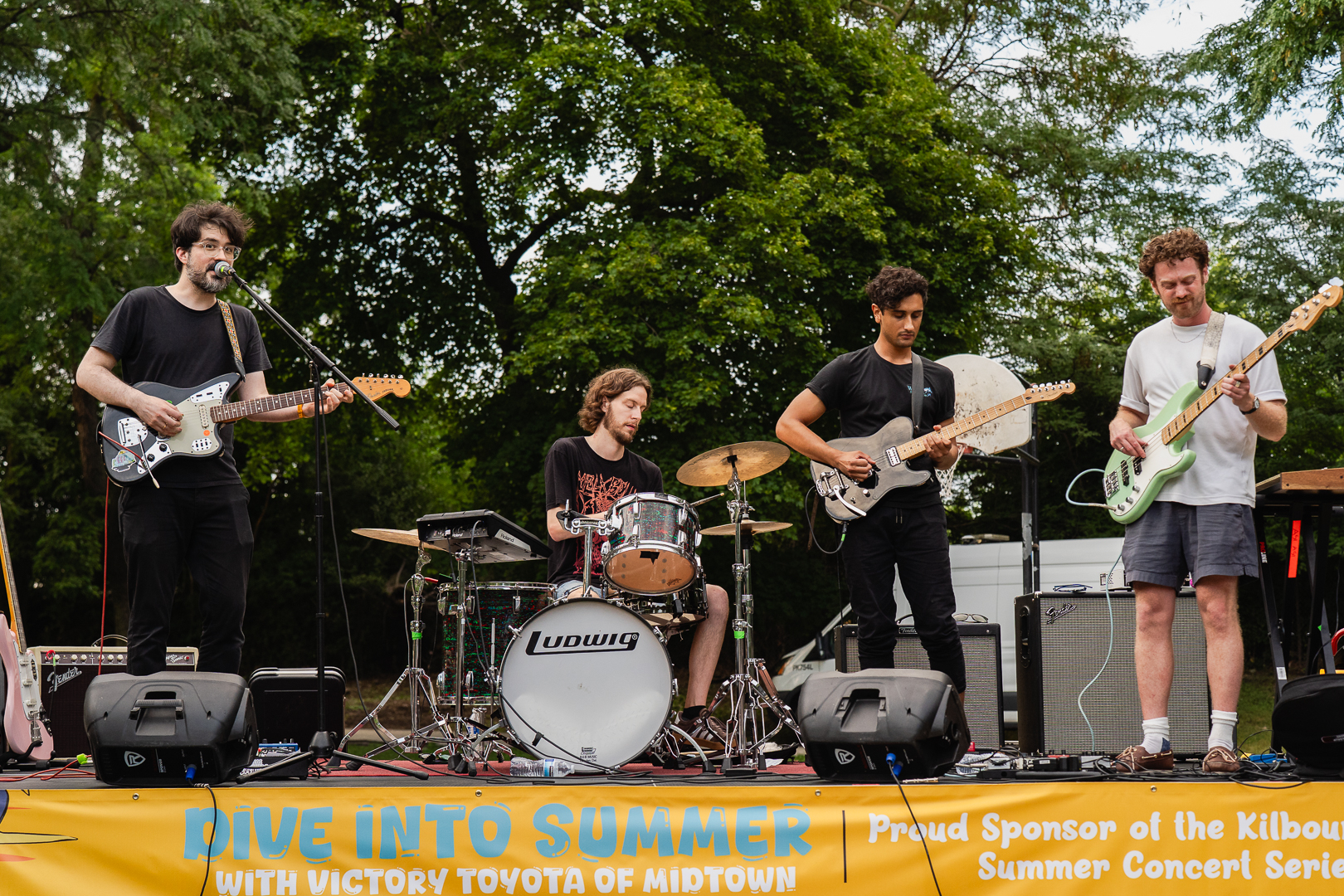 Four musicians play on an outdoor stage backed by trees.