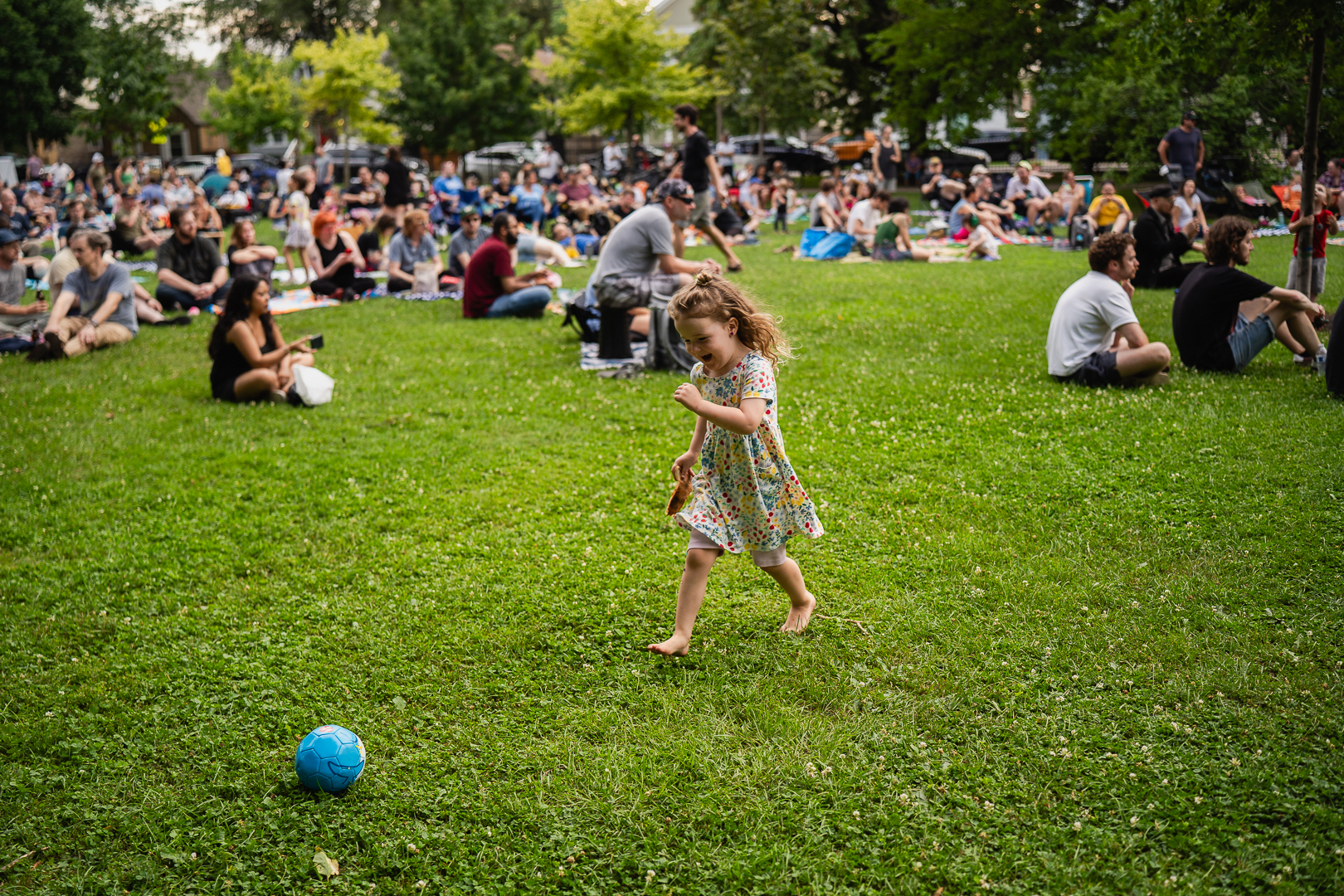 A barefoot toddler runs across a grassy field towards a small blue soccer ball while a crowd sits in the background.