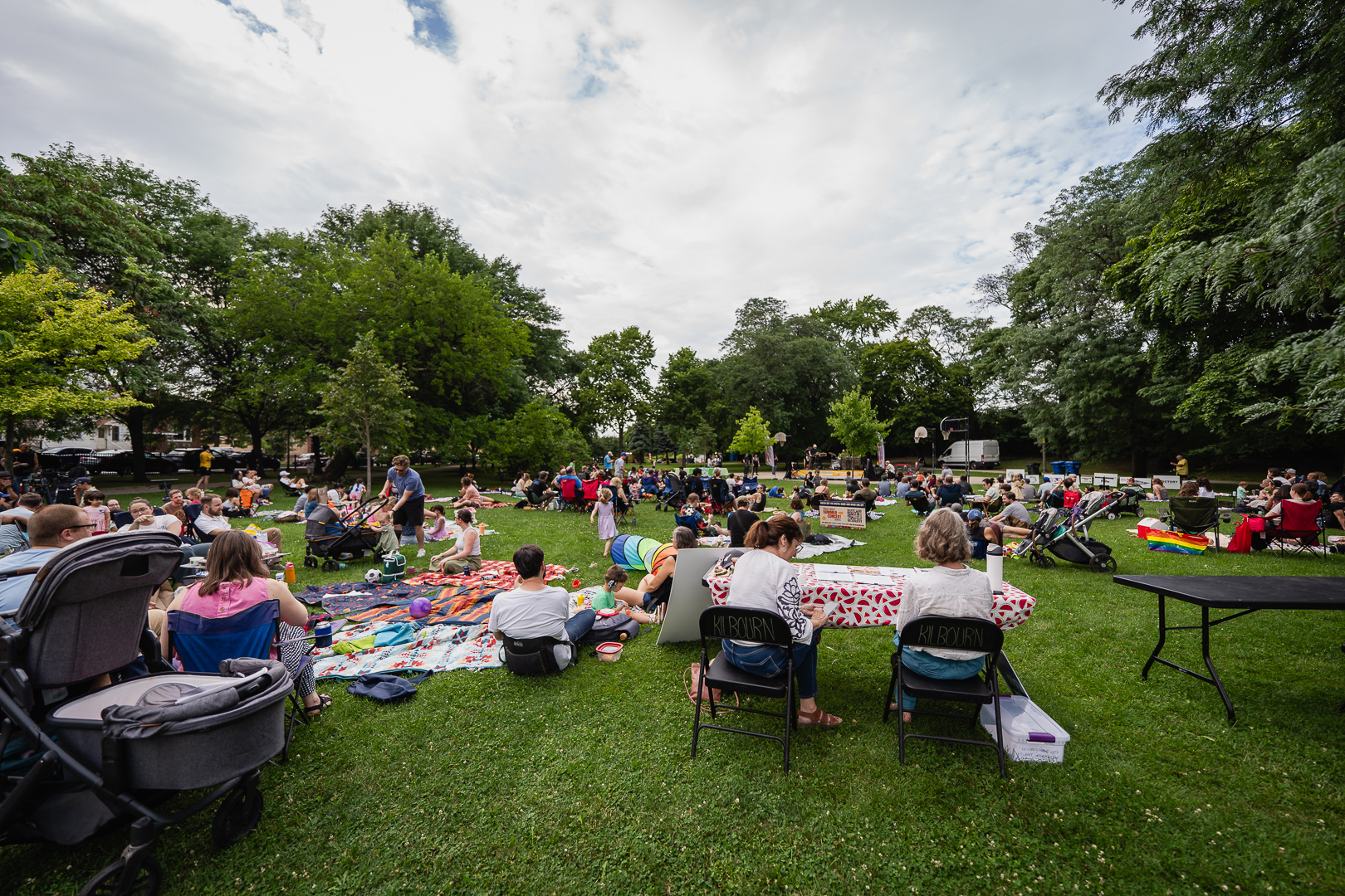 Families gather on blankets and lawn chairs at an outdoor event in a park.
