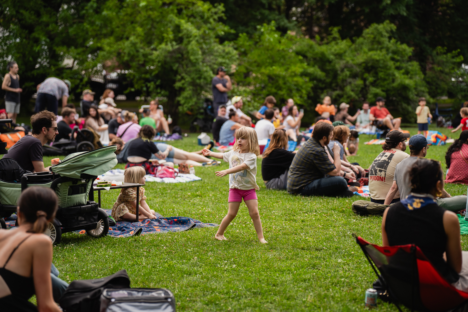Toddler dances on grass at a park, surrounded by seated people.