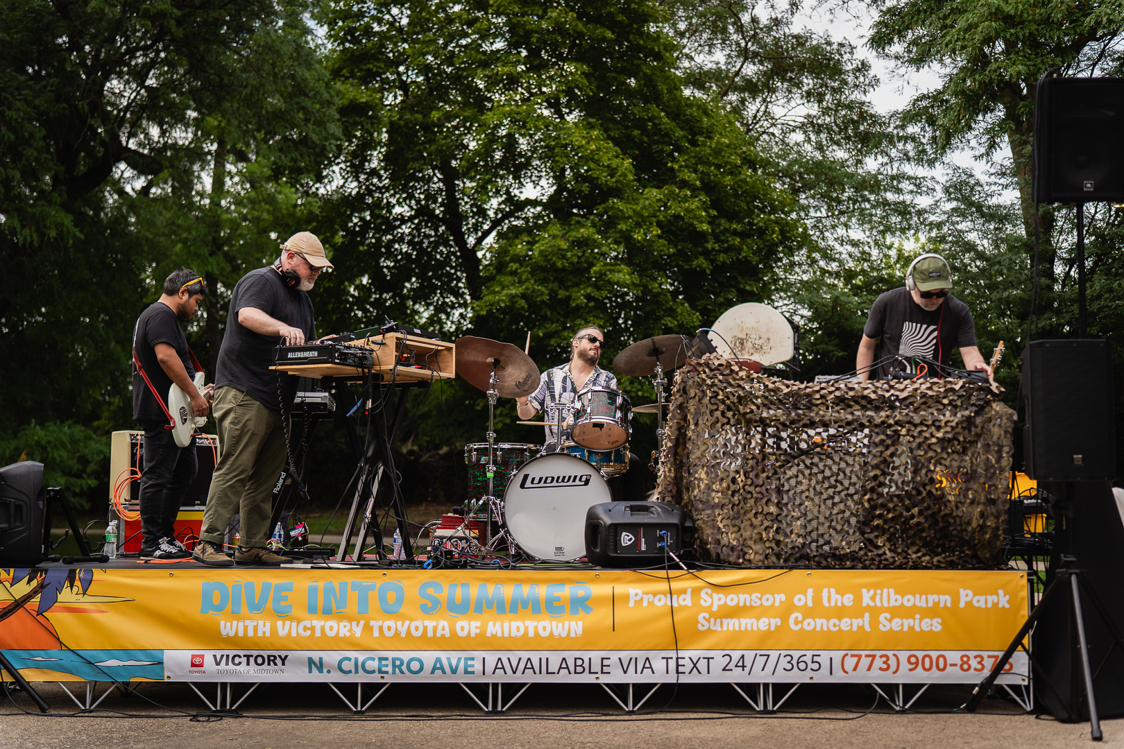 A band plays on an outdoor stage backed by trees.