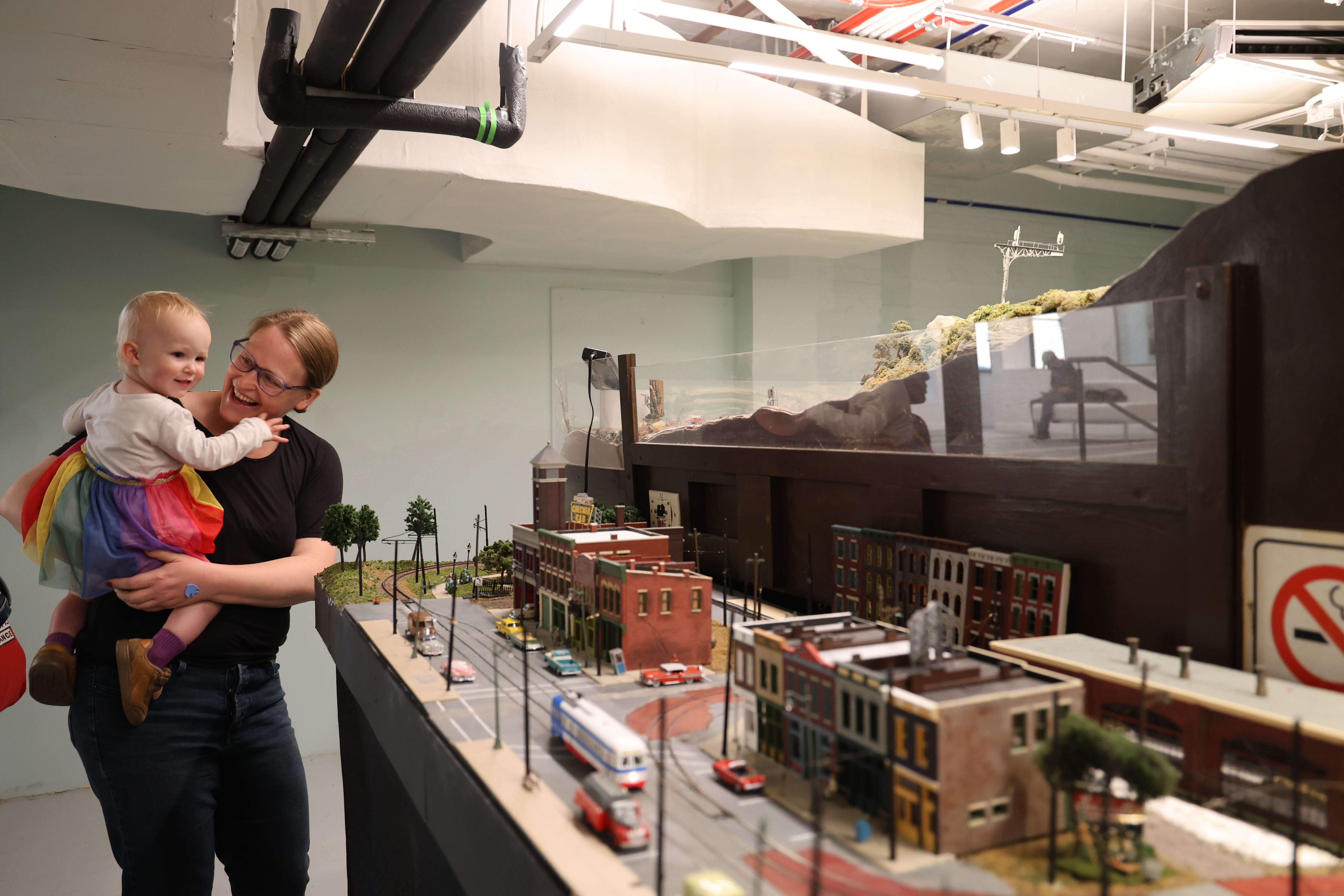Adult and toddler look at a detailed model town with miniature buildings and vehicles.