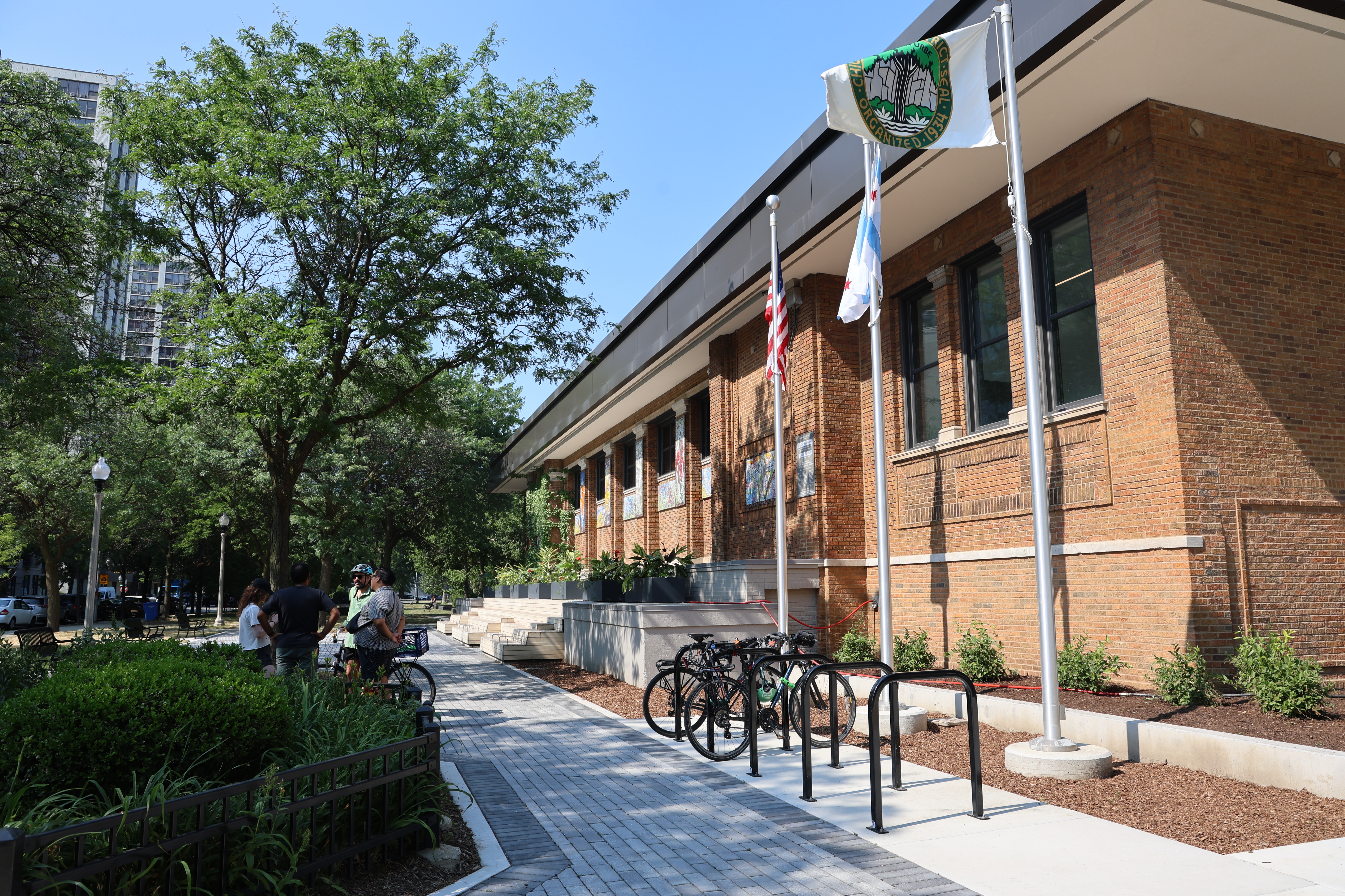 Brick building with flags, bike rack, and people on a paved path. Trees and a park are visible.