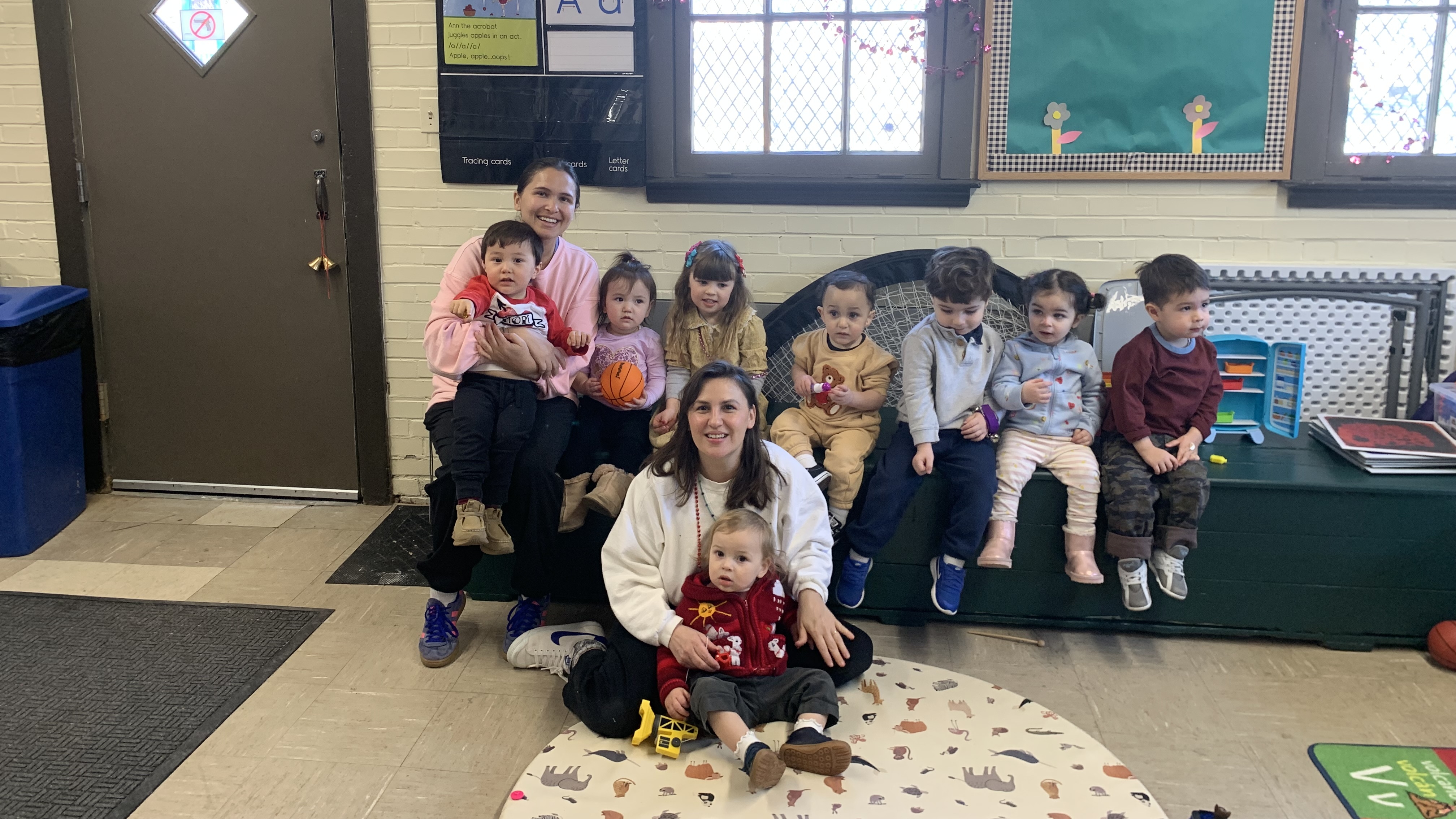 Two smiling women with eight young children in a brightly lit classroom.