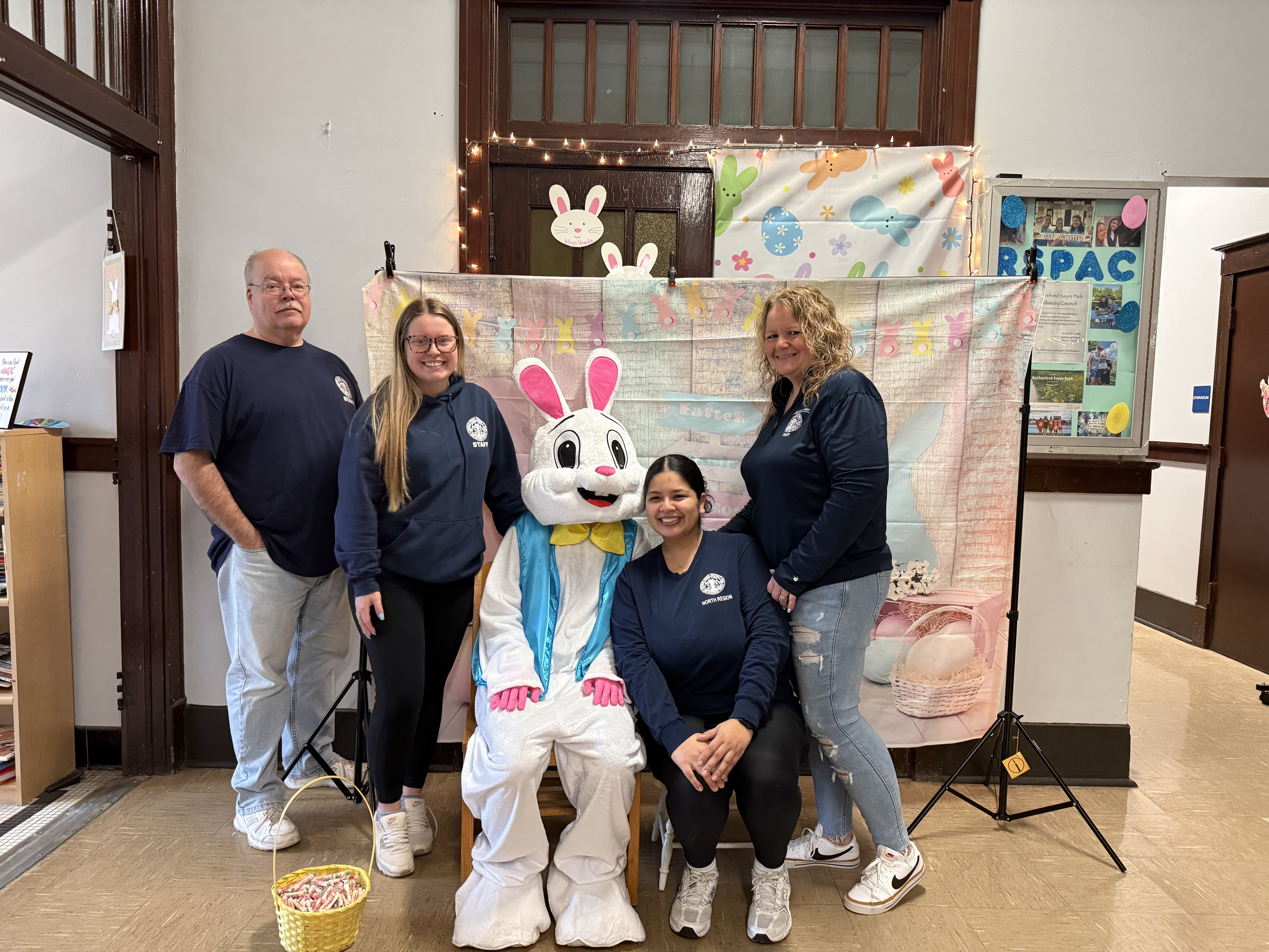 Four people and an Easter Bunny mascot pose for a photo in front of an Easter-themed backdrop.