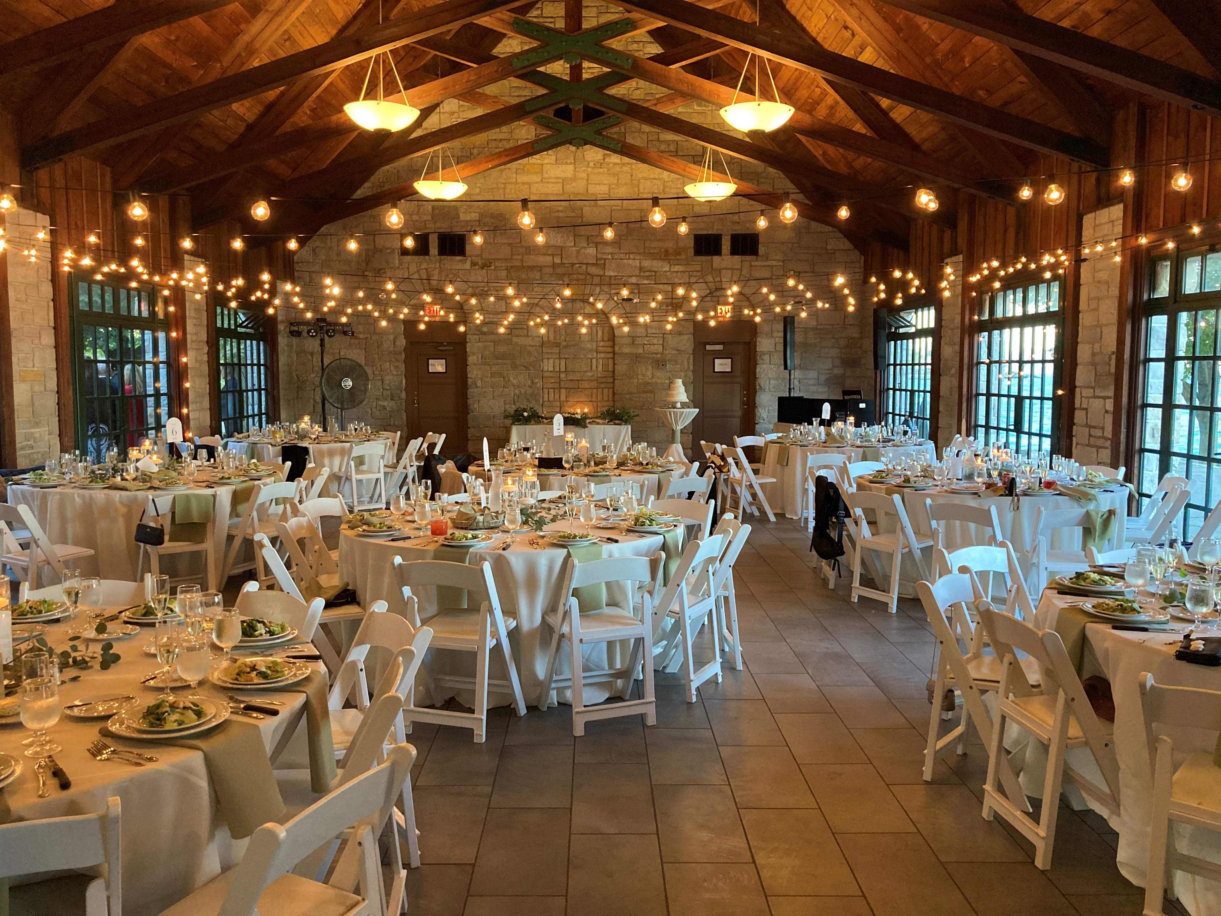 Rustic wedding reception hall with round tables, white folding chairs, and string lights.