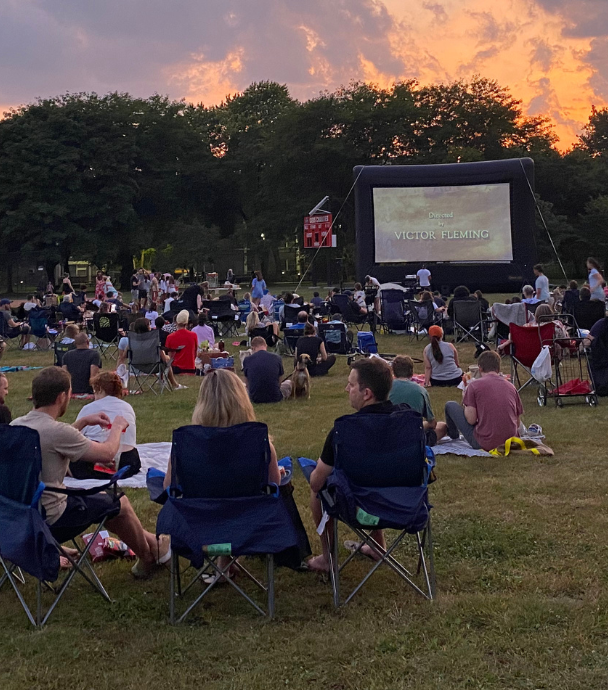 People sit in lawn chairs watching a movie on an inflatable outdoor screen at sunset.