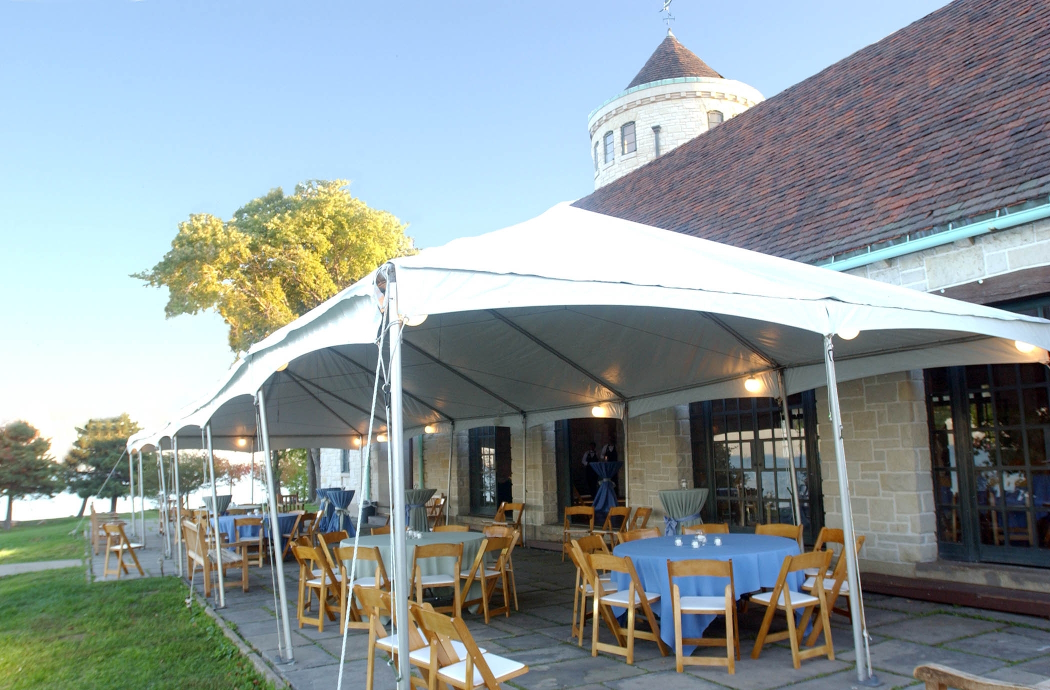 Outdoor event tent set up next to a stone building with round tables and chairs.