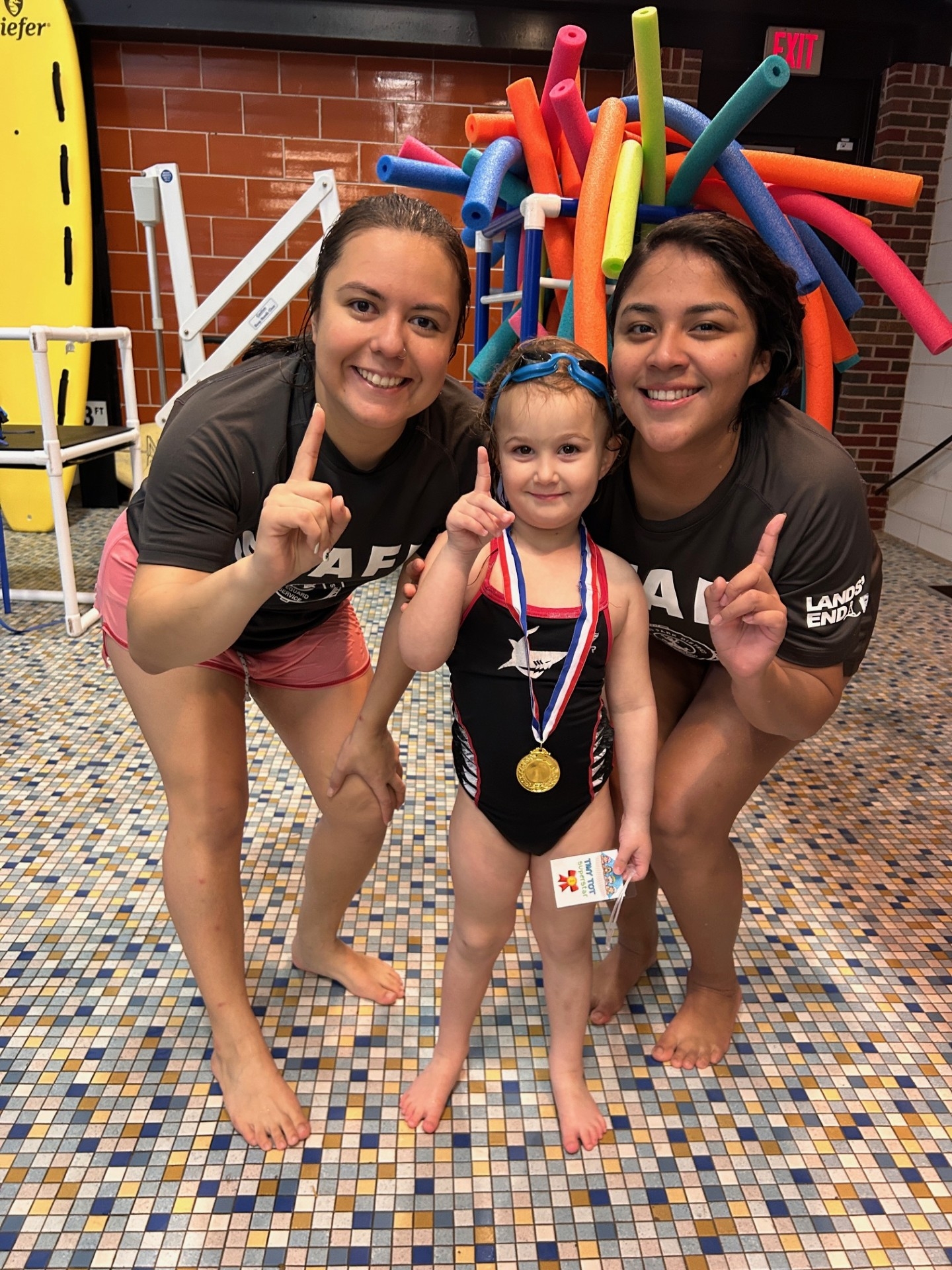 A young swimmer wearing a medal and swim ribbon poses with two swim instructors by the pool. They each hold up one finger.