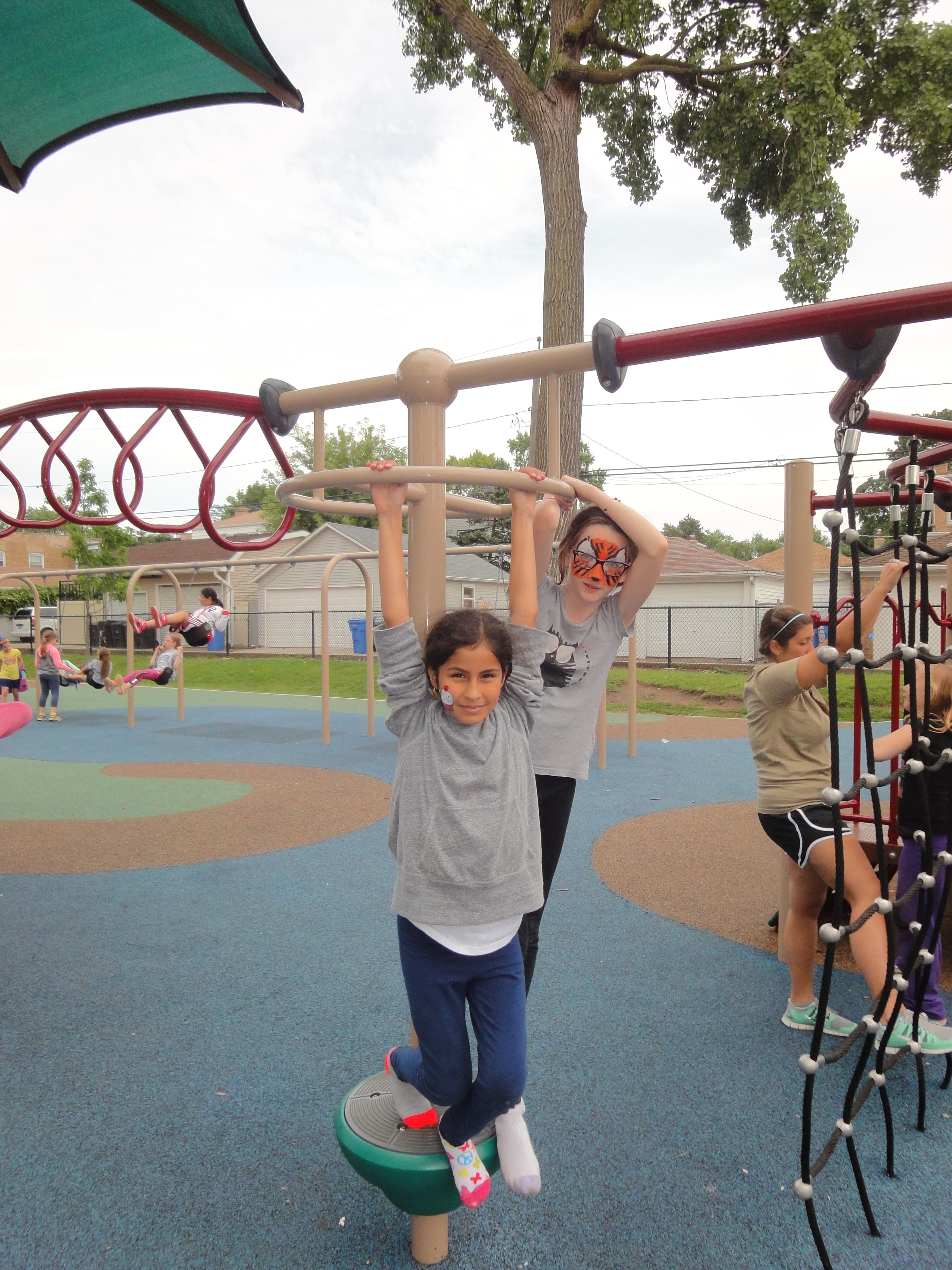Two children play on a playground spinner. Other children play on equipment and swings in the background.