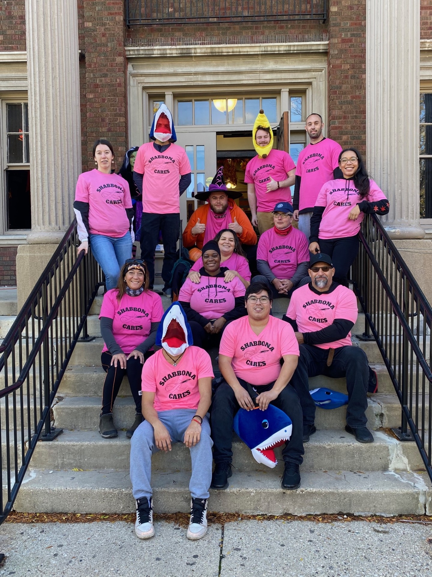 Group photo of people wearing pink Shabbona Cares t-shirts on steps of brick building. Some are wearing playful costumes.
