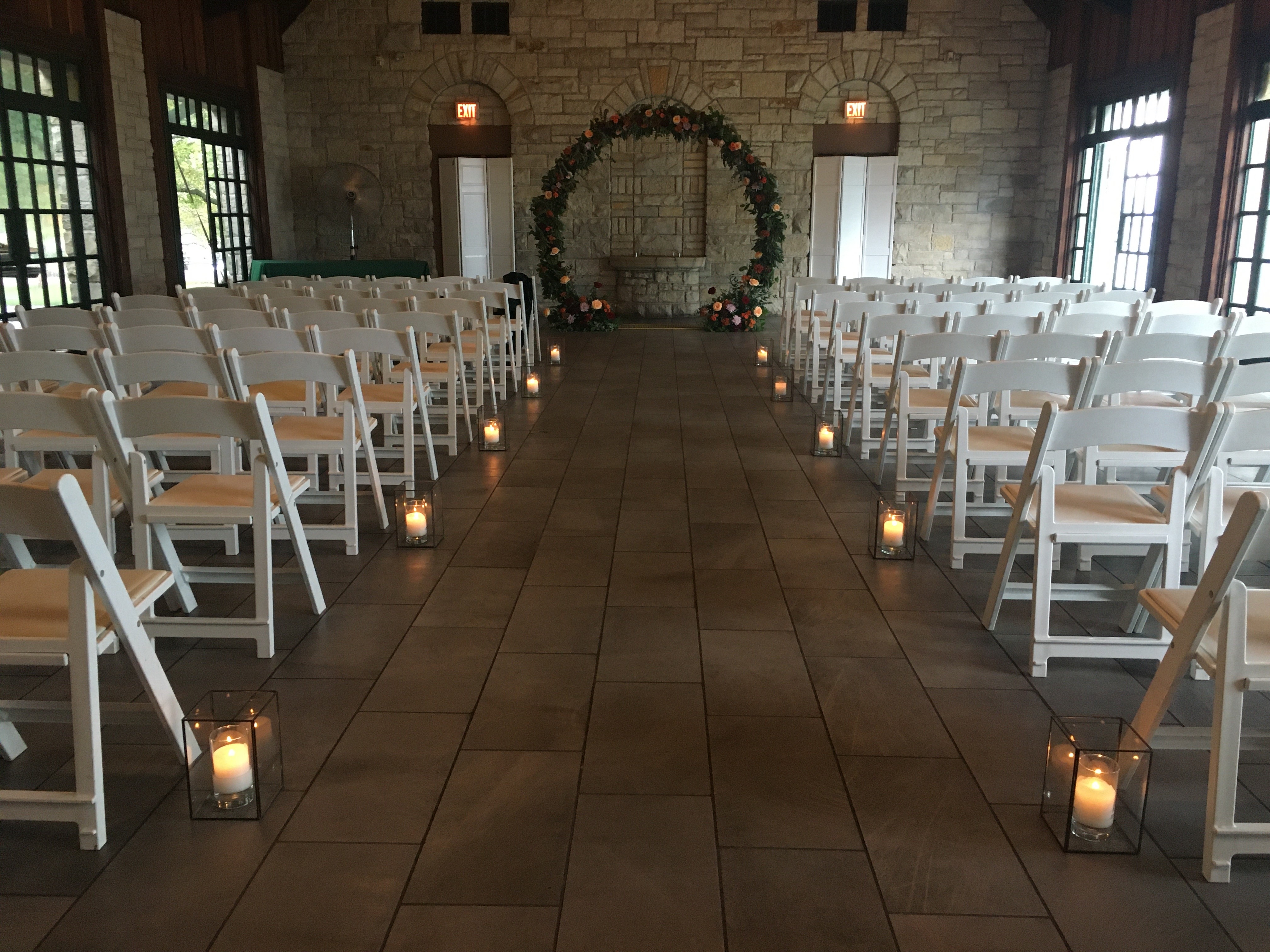 Indoor wedding ceremony setup with white chairs, floral arch, and candlelit aisle.