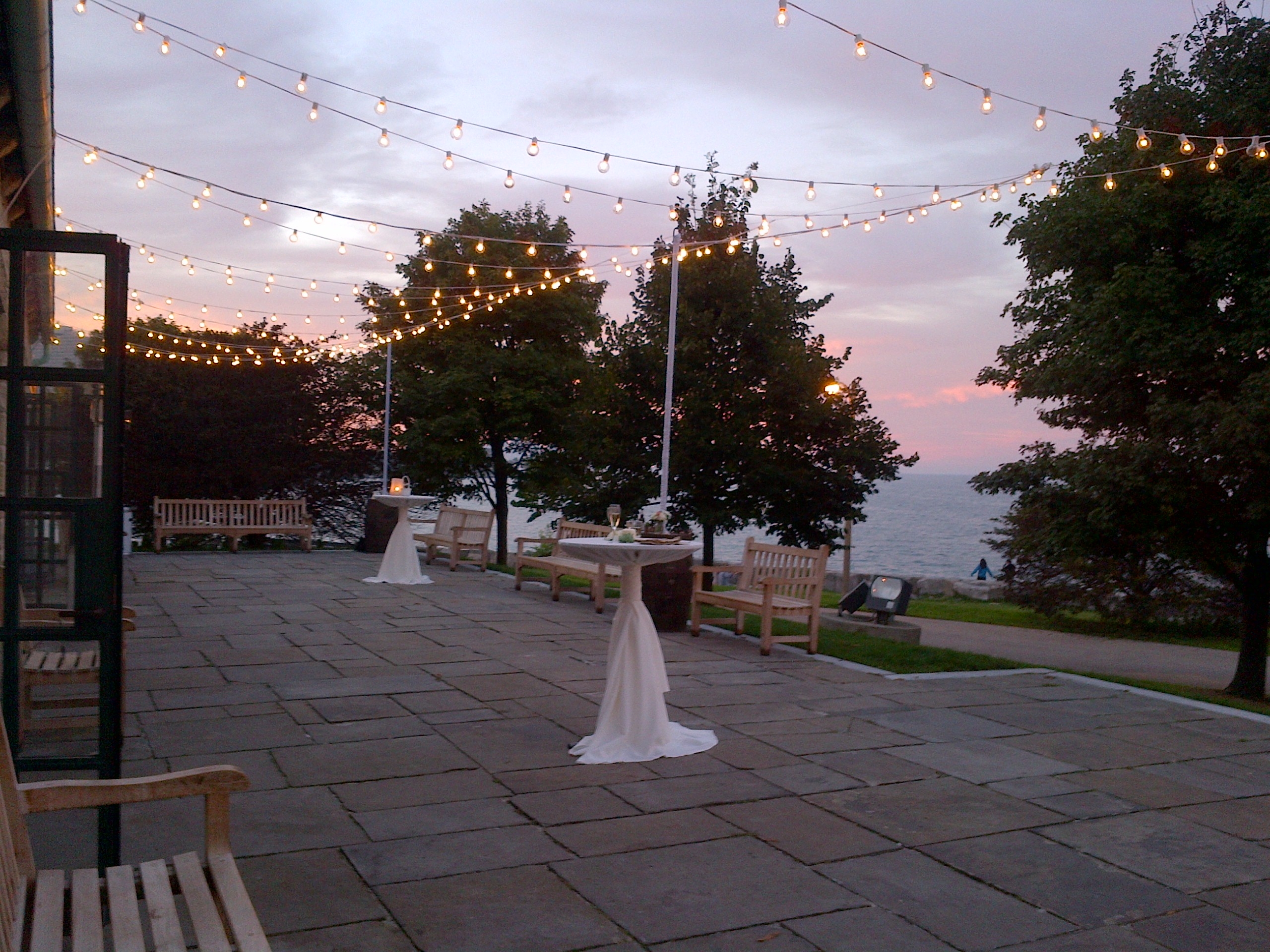 Outdoor patio with string lights at sunset. Tables and benches. Water visible in the background.
