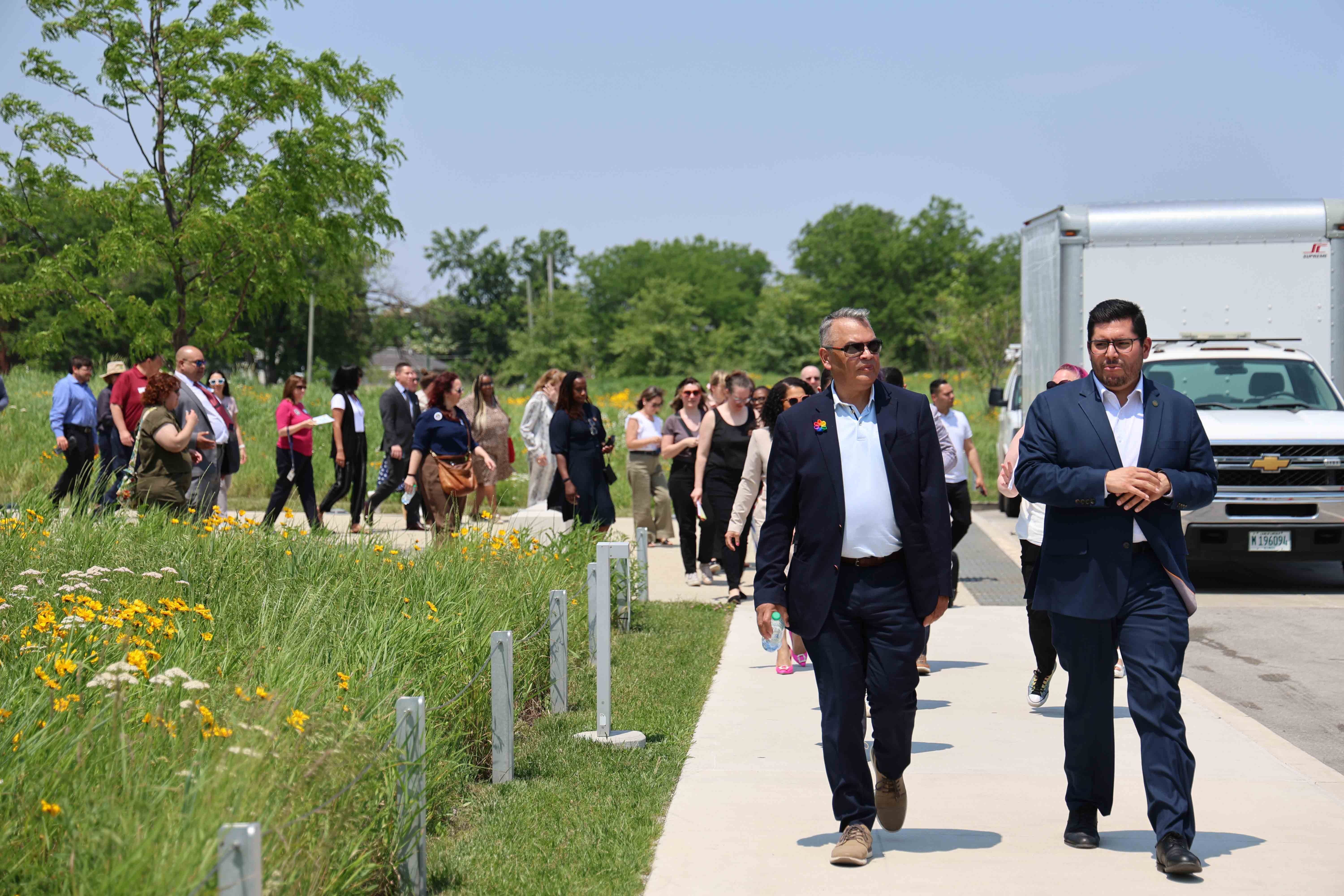 A group walks along a paved path through a field of wildflowers. Two men in suits lead the group.