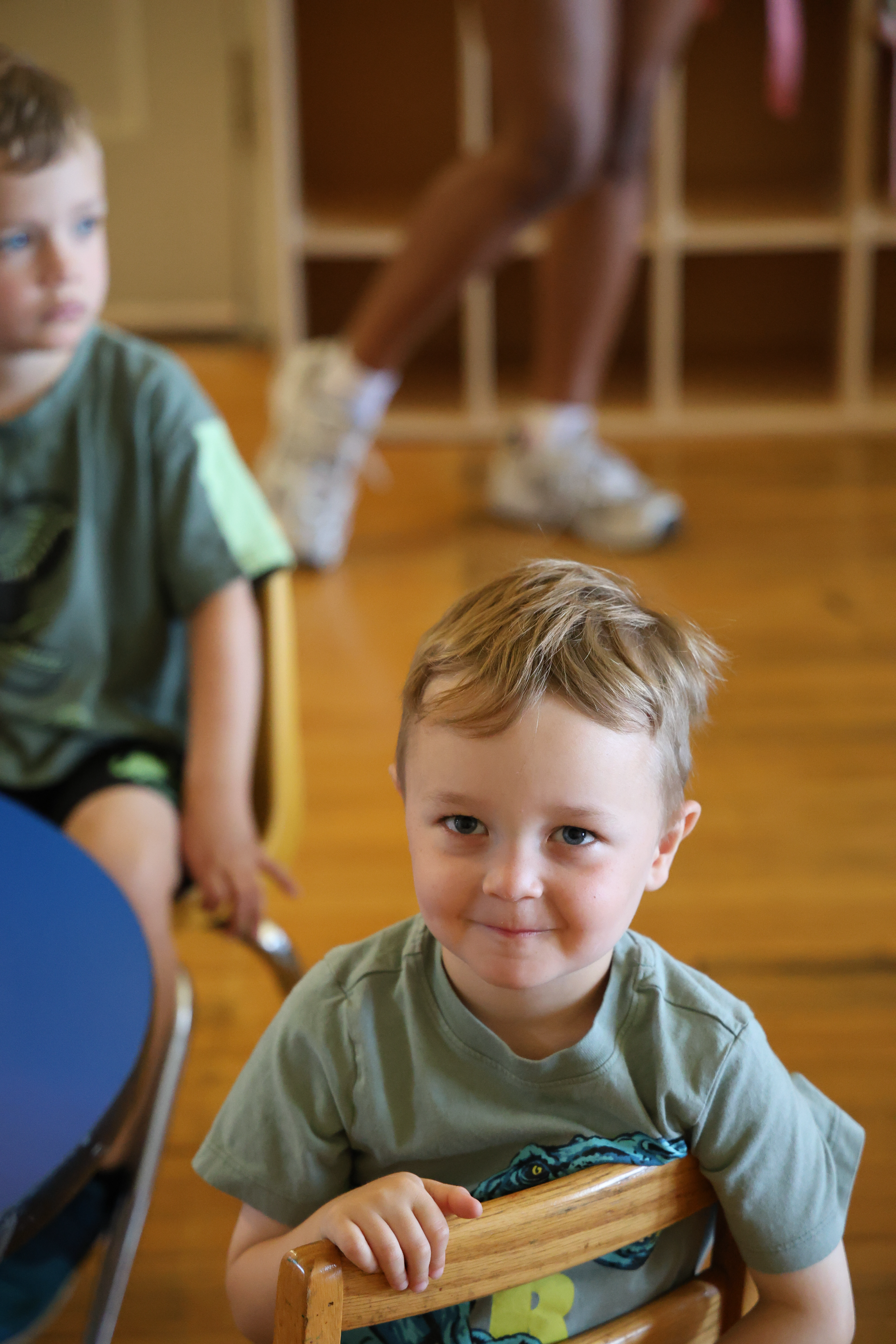 Smiling blond boy in a green t-shirt sits in a small wooden chair.