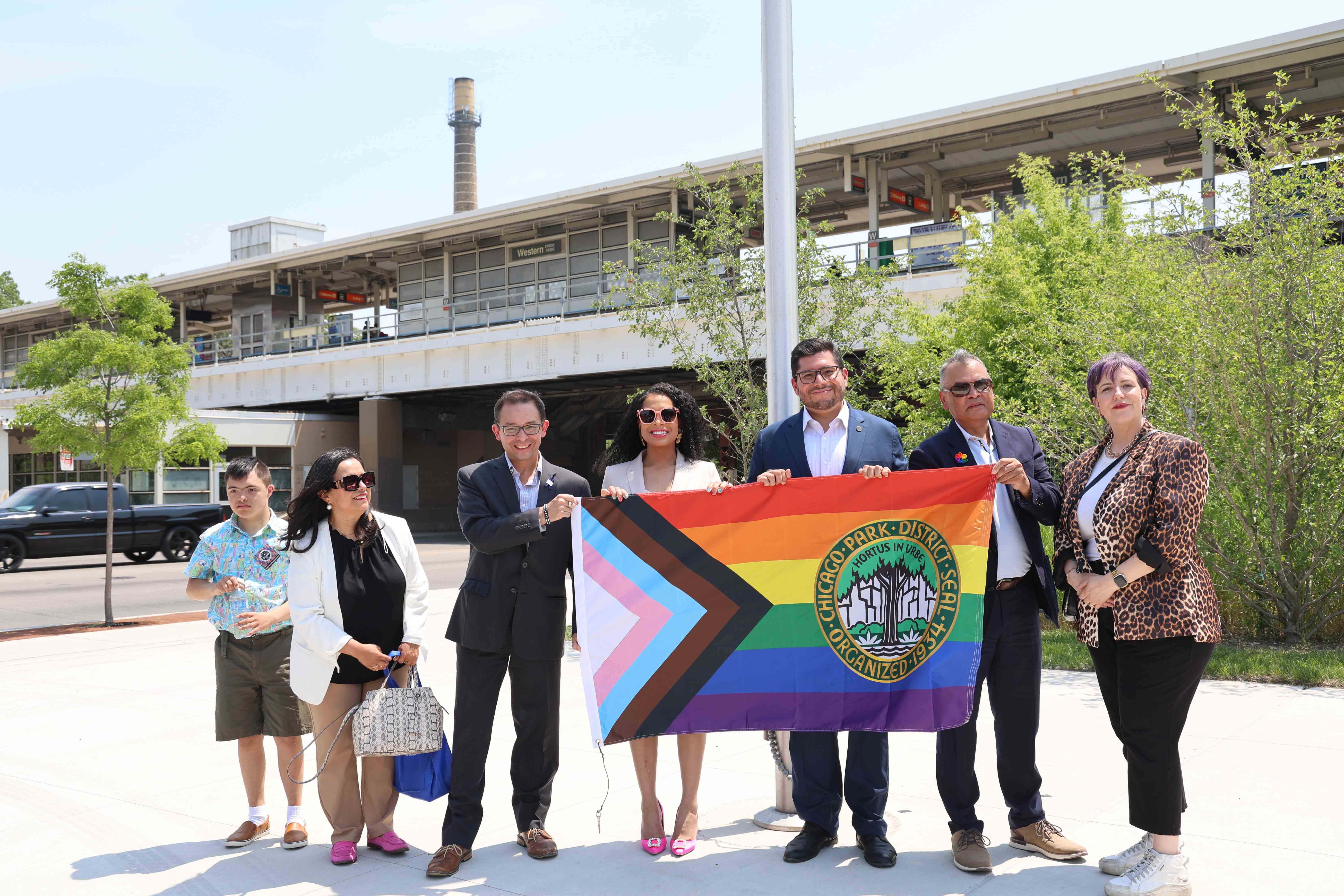 Group holds Progress Pride flag with 첥Ƶ logo beneath elevated train platform.