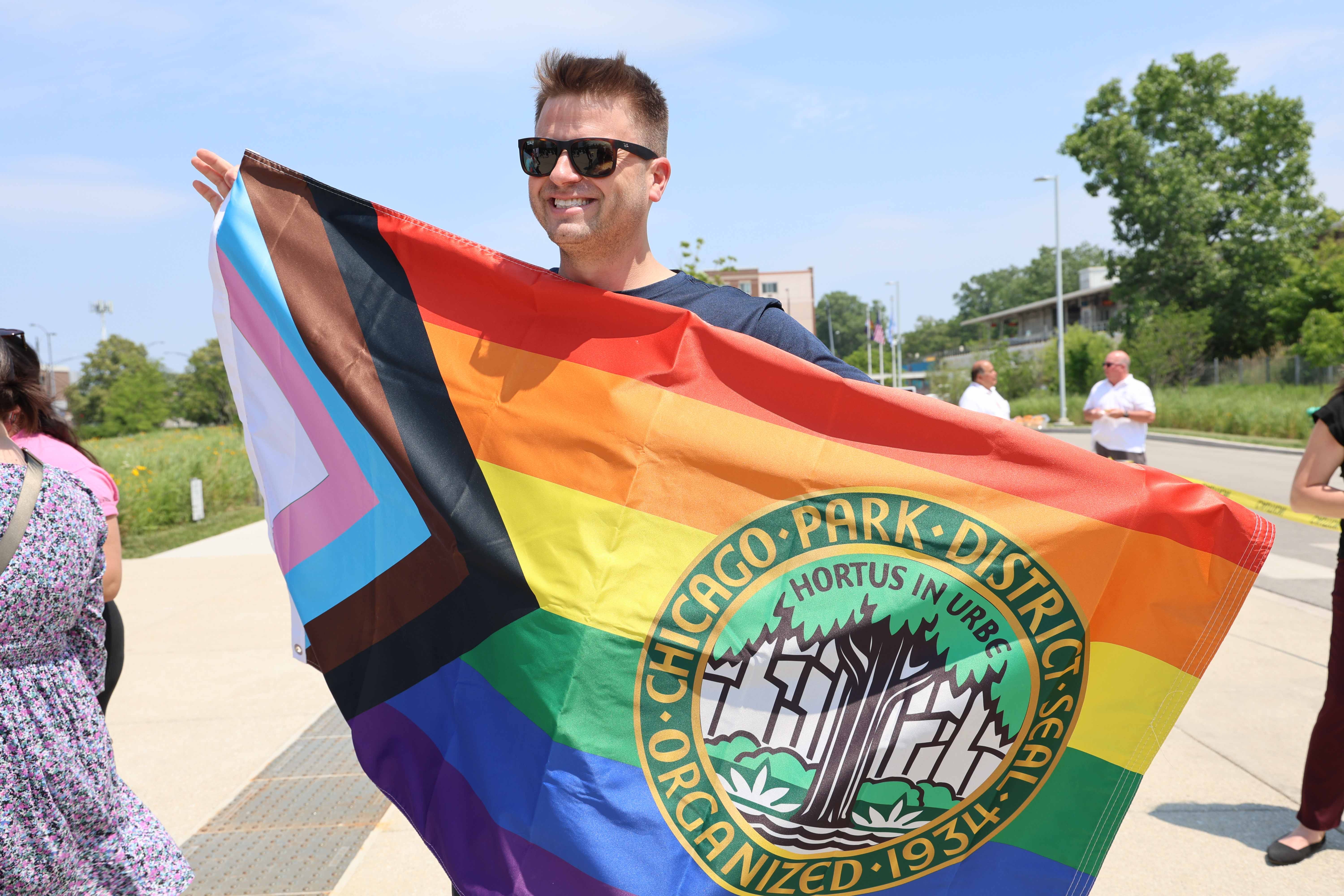Smiling person holding a Progress Pride flag with the 첥Ƶ logo.