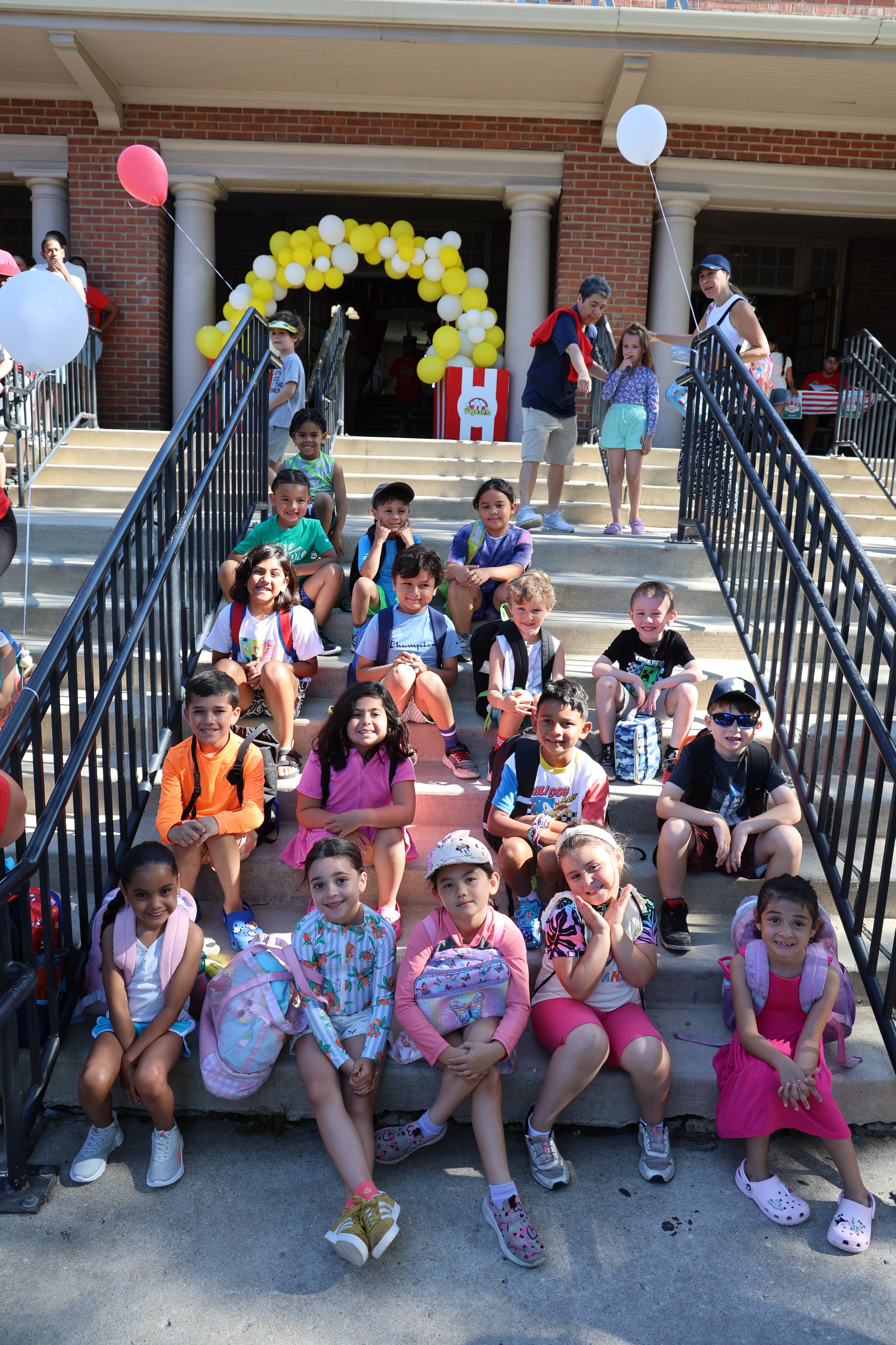 Children with backpacks sit on steps outside a brick building decorated with balloons.