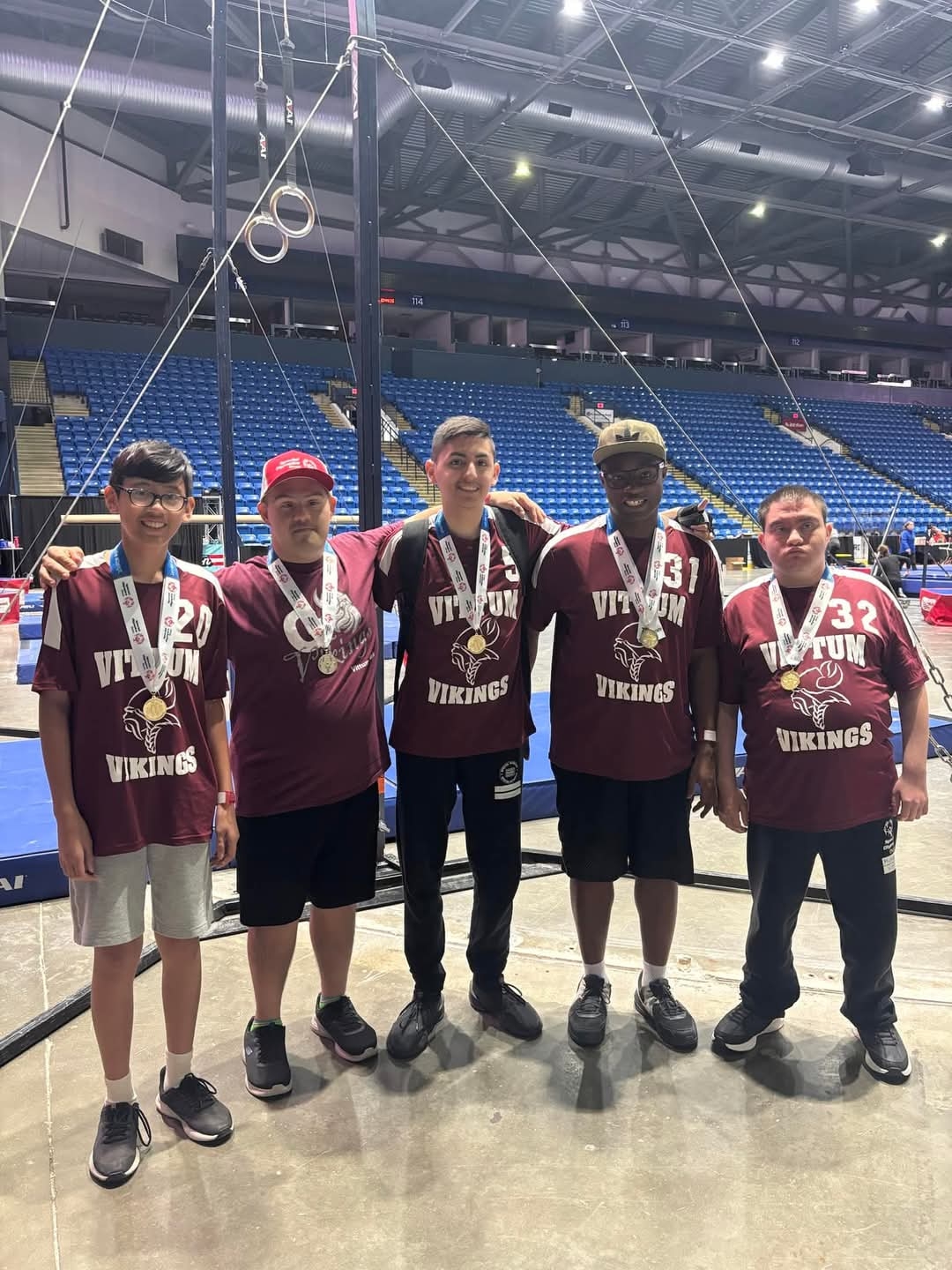Five athletes wearing Vikings team shirts and medals pose together at a gymnastics competition.