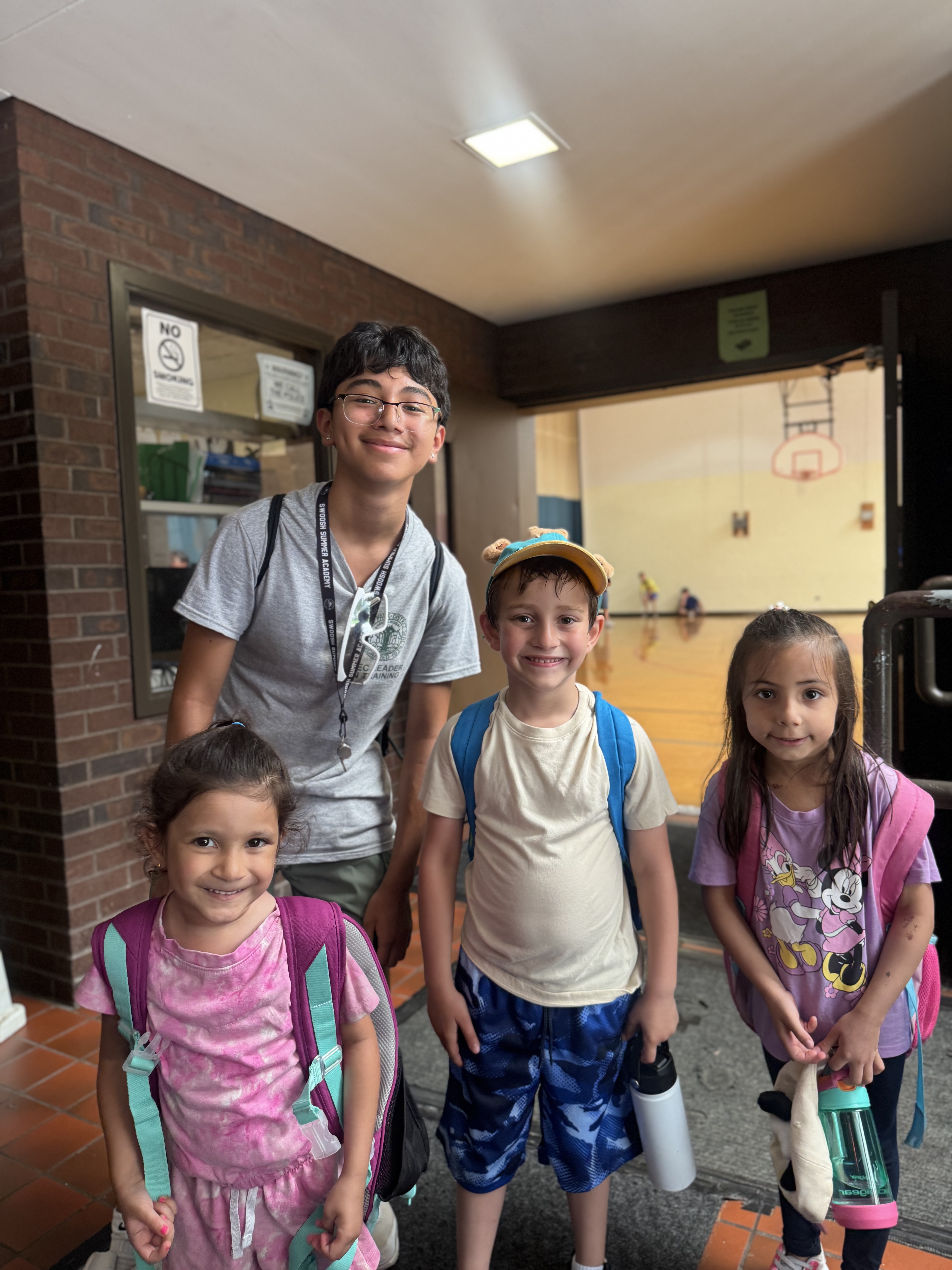 Four children with backpacks stand outside a gym.