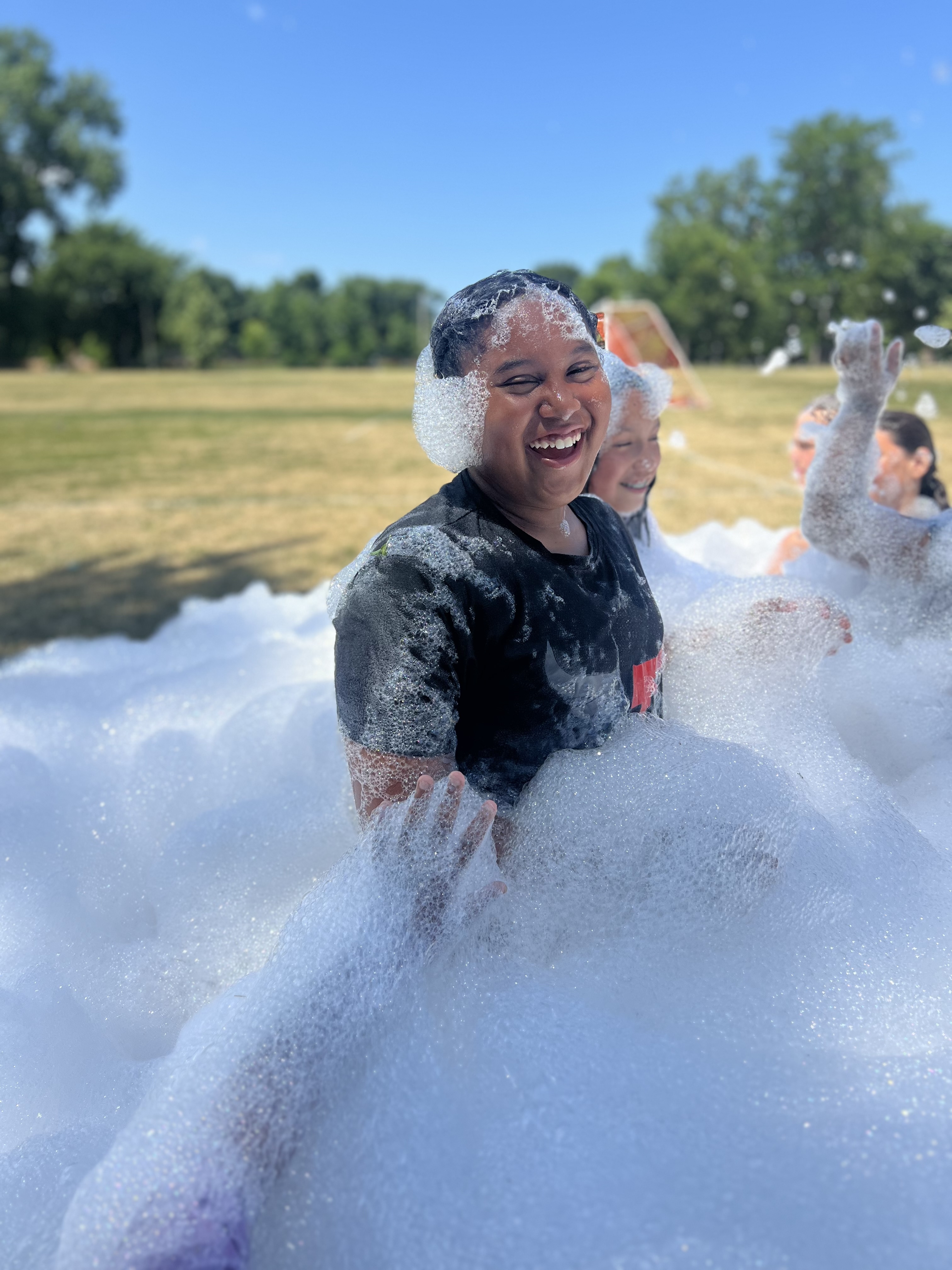 A laughing child sits in a mound of white foam at an outdoor event.