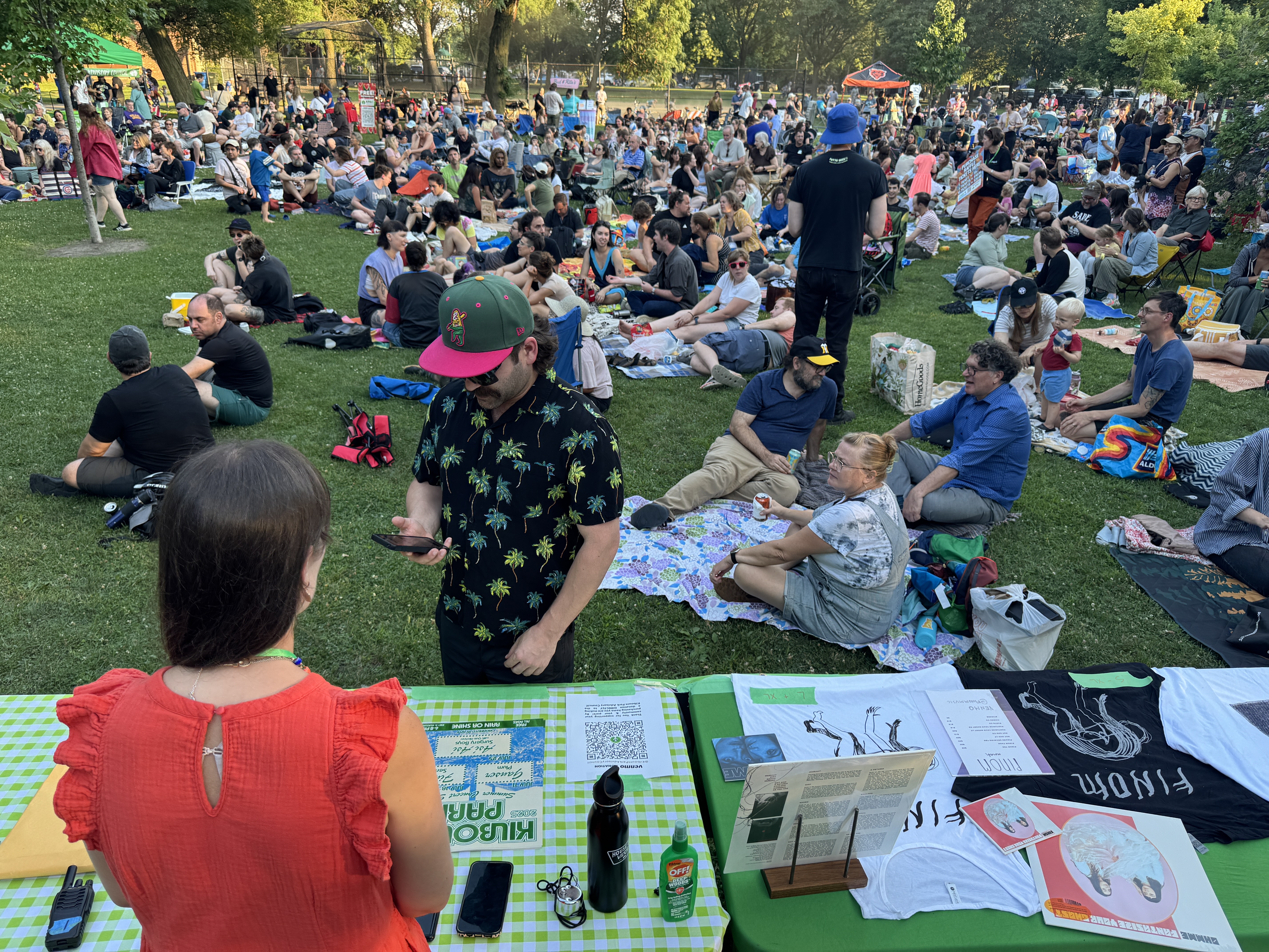 Crowd sits on grass at outdoor concert, vendor table in foreground.
