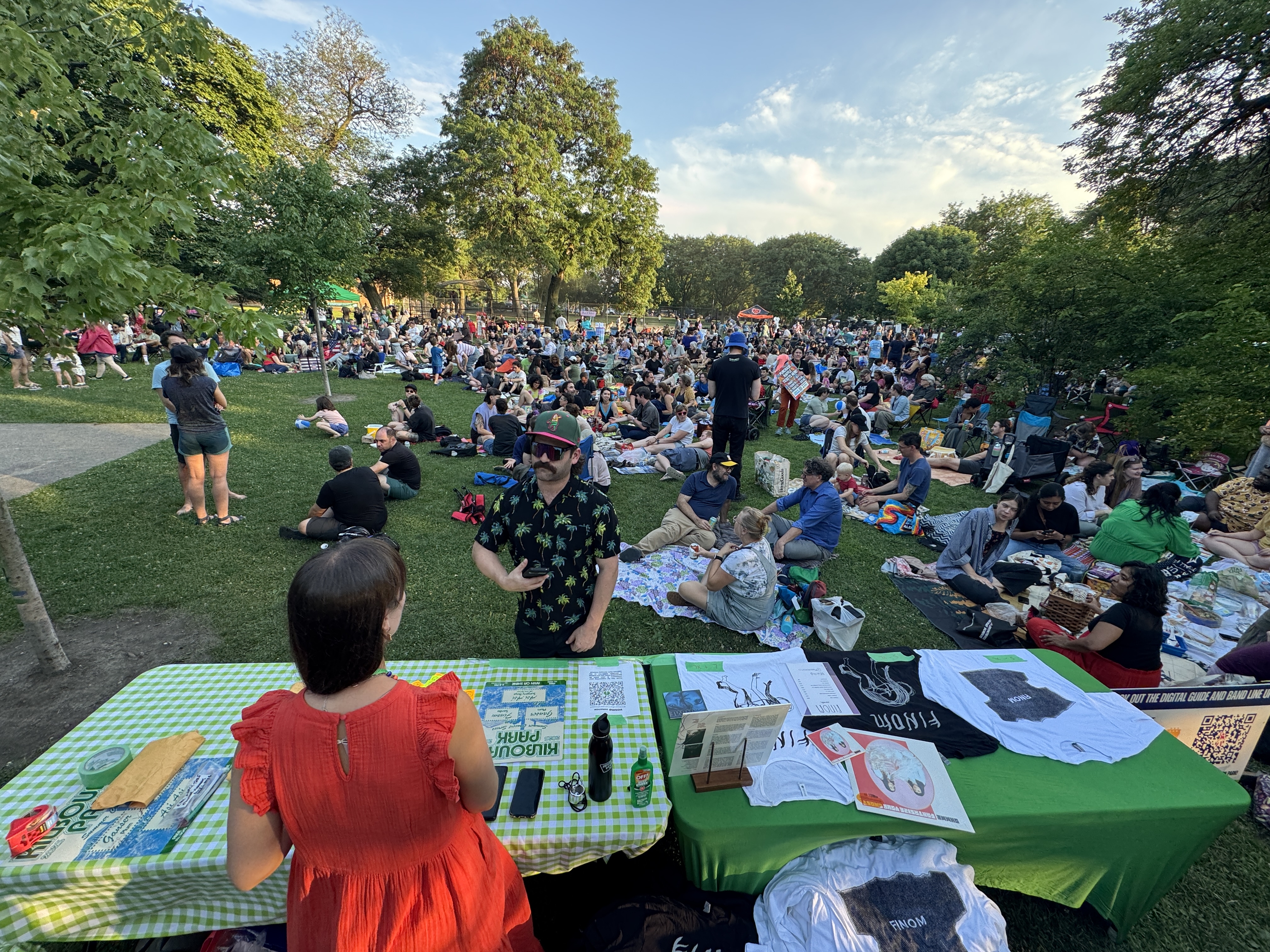 Crowd sits on lawn at outdoor concert, vendor tables in foreground.