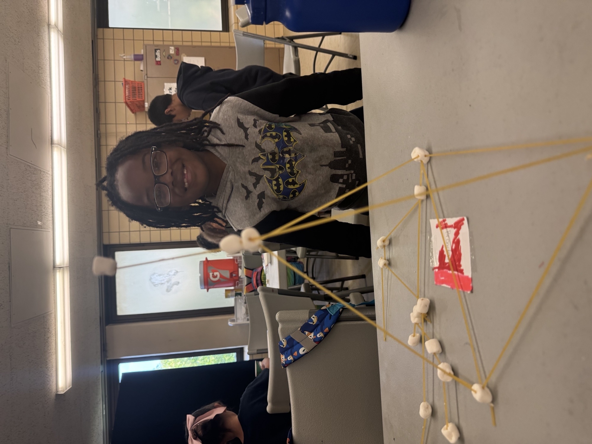 A student with dreadlocks smiles near a spaghetti and marshmallow structure.