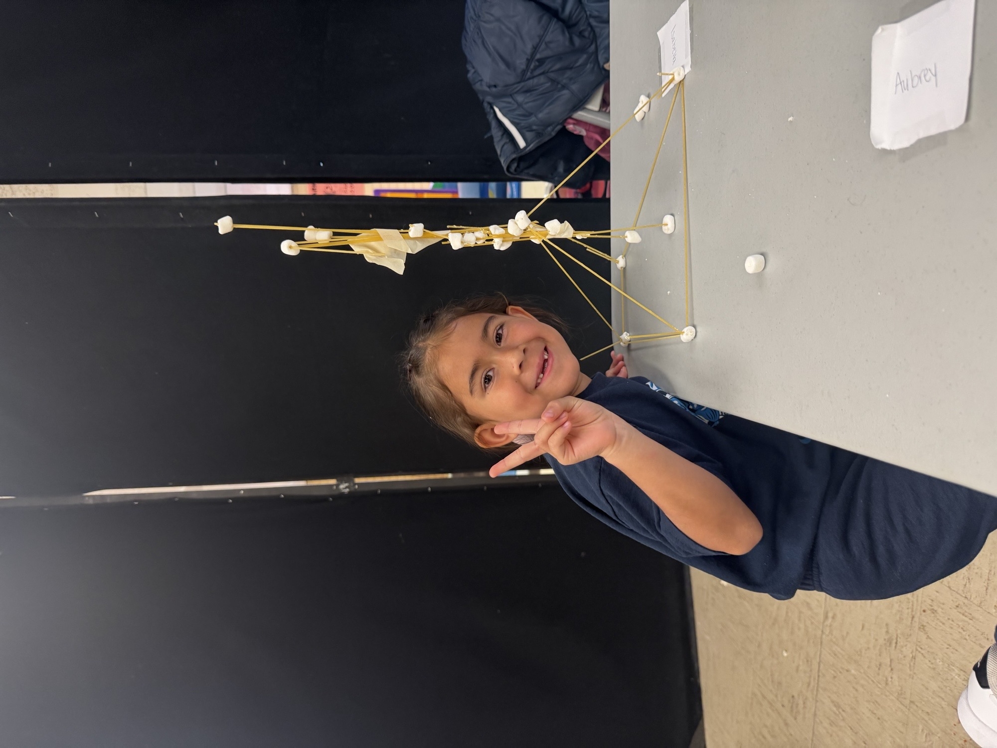 Smiling child with spaghetti and marshmallow tower, peace sign.