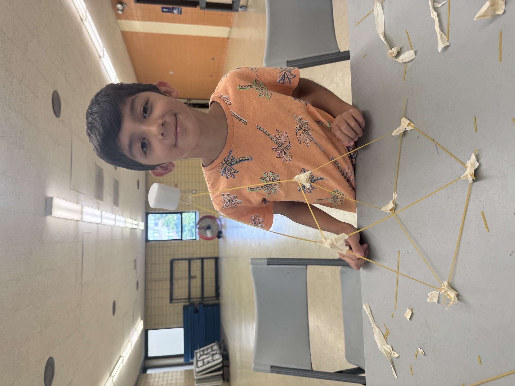 A child smiles by a table with a spaghetti and marshmallow structure.