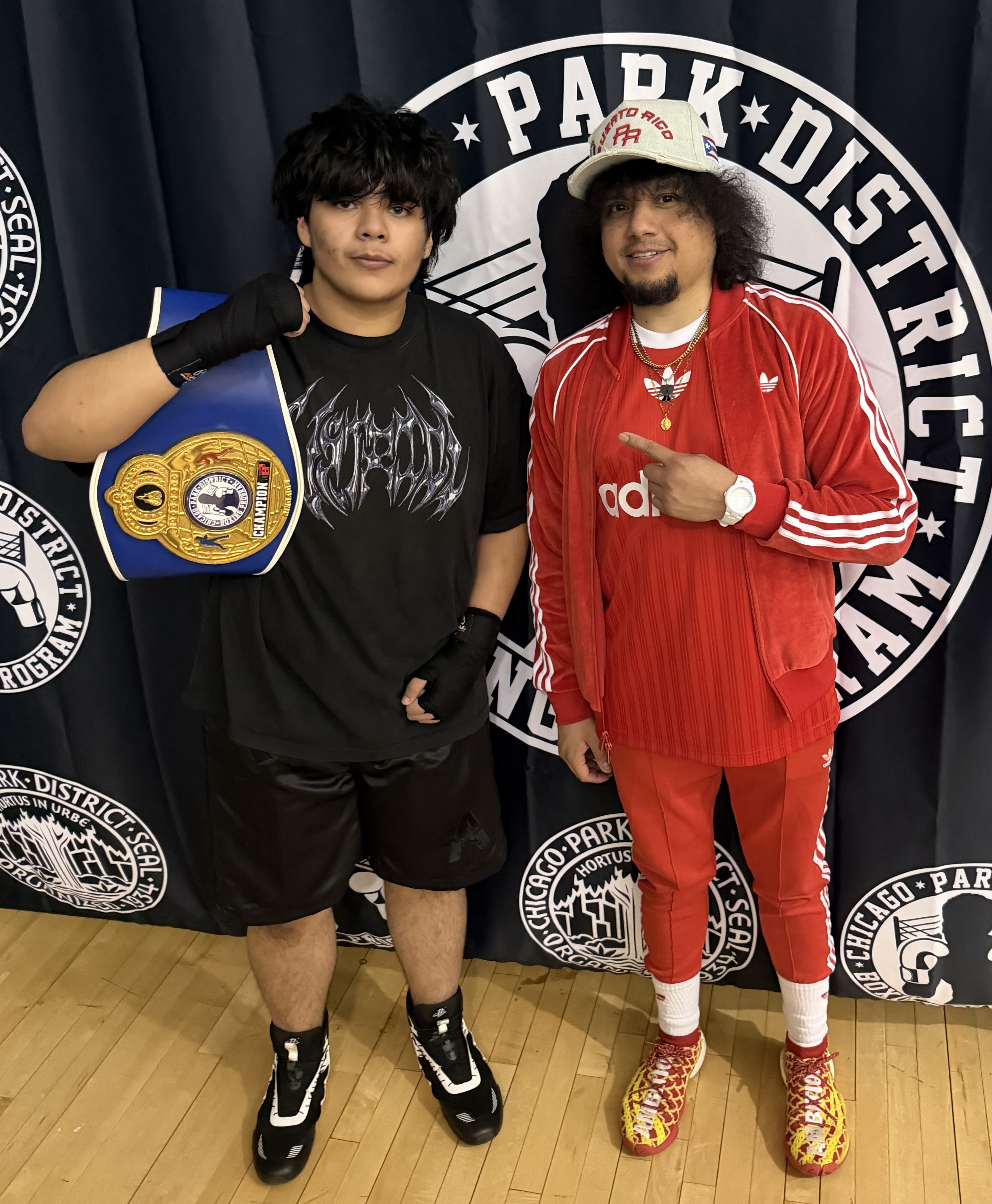 Two men pose; one holds a boxing champion belt, the other wears a red Adidas tracksuit.